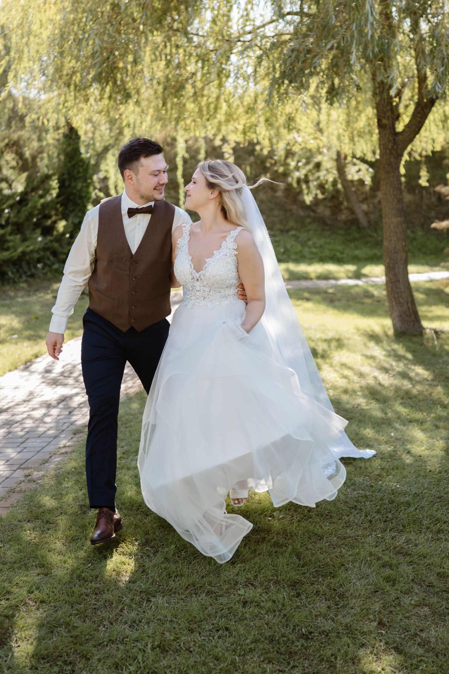 Couple walking together through a garden setting with willow trees, the bride's white dress flowing in the sunlight.