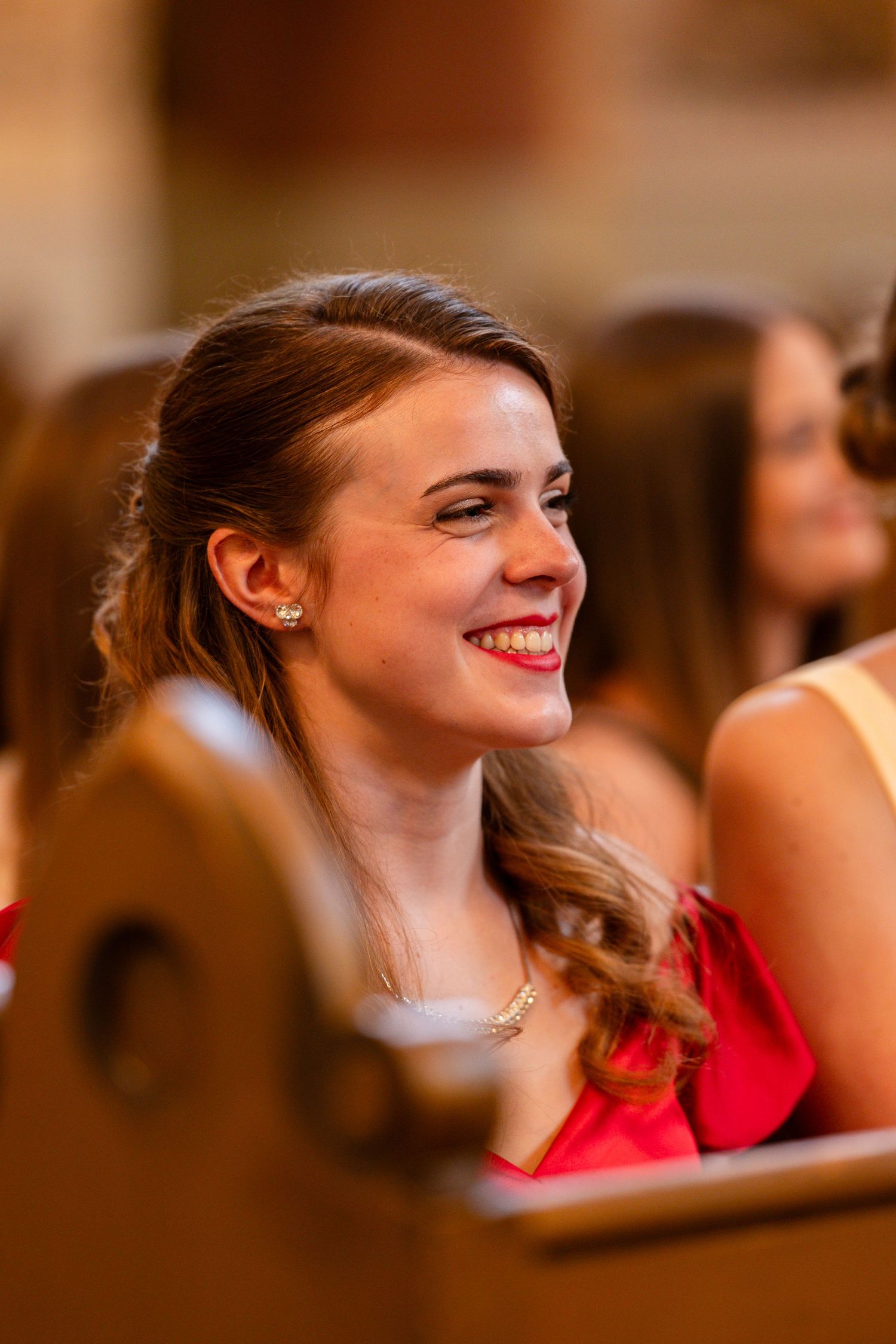 Guest in a red dress laughs during wedding ceremony in church pews.