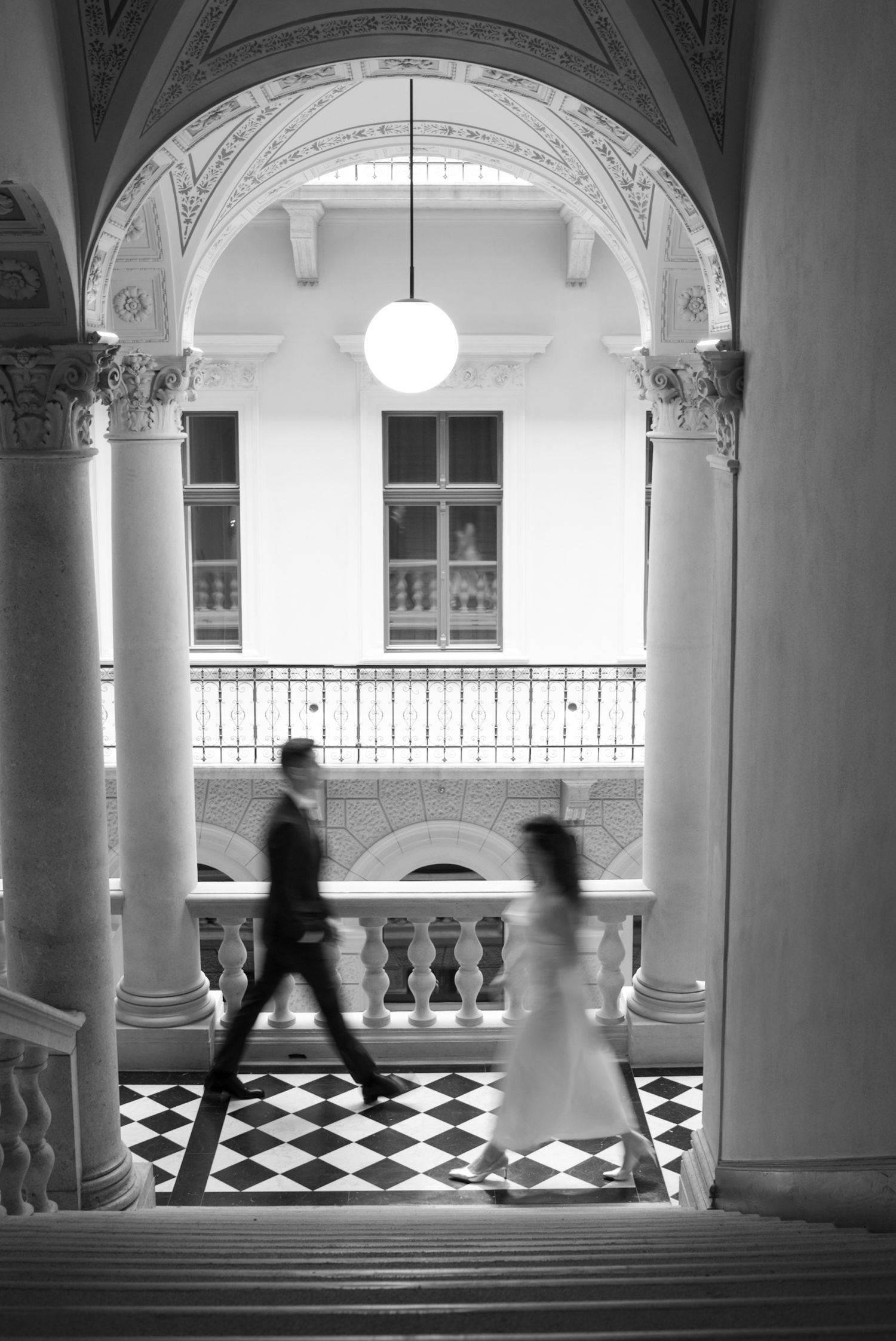 Blurred motion of bride and groom walking through elegant black and white checkered hall with arched columns.