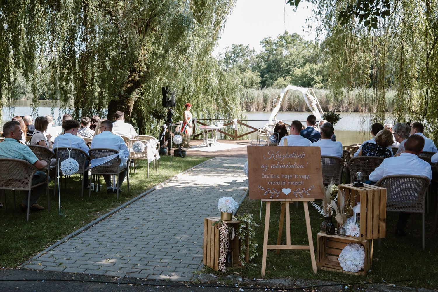 Outdoor wedding ceremony setup with white chairs and wooden decor under willow trees.