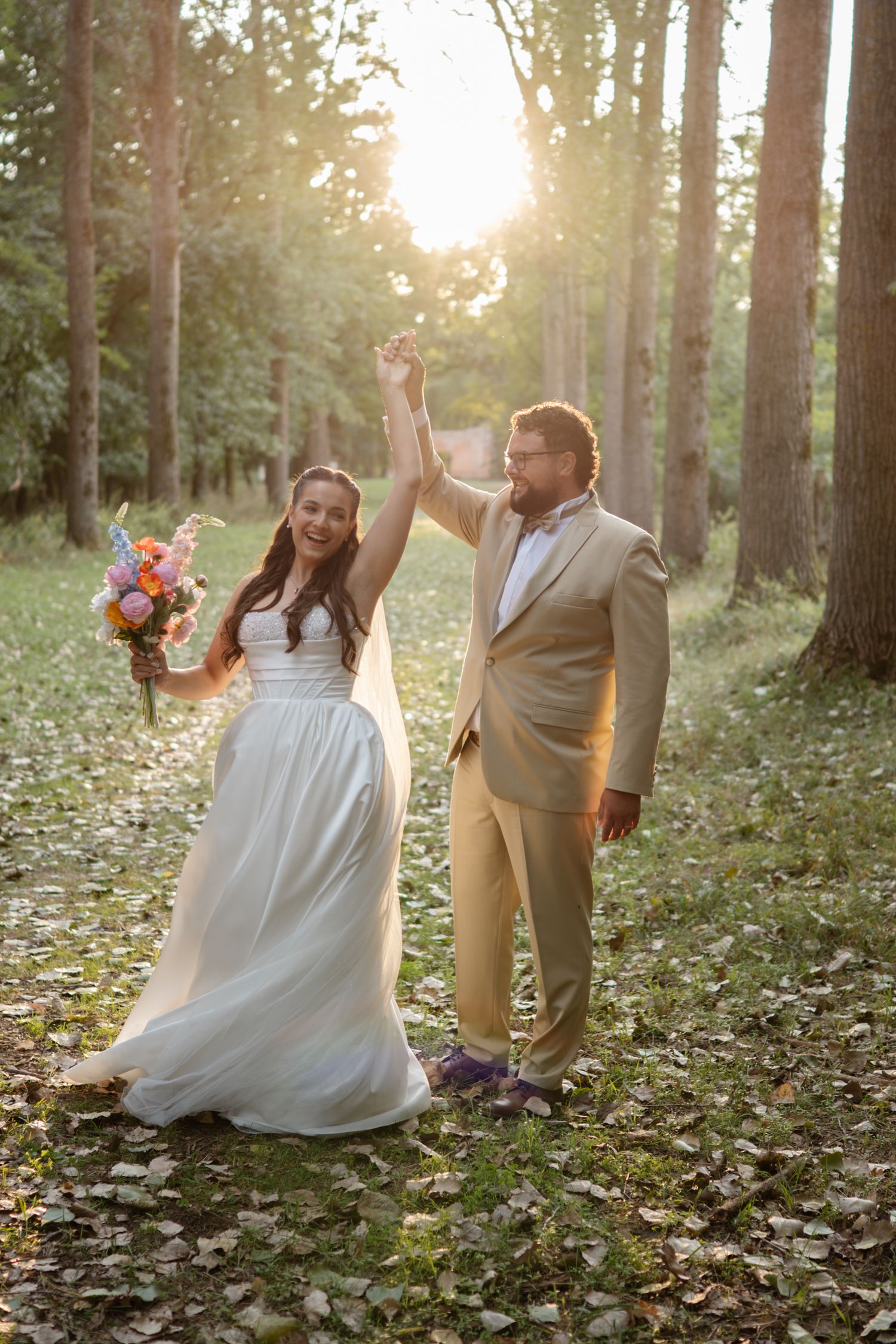 A couple in wedding attire dances playfully in a sunlit forest with fallen leaves on the ground.