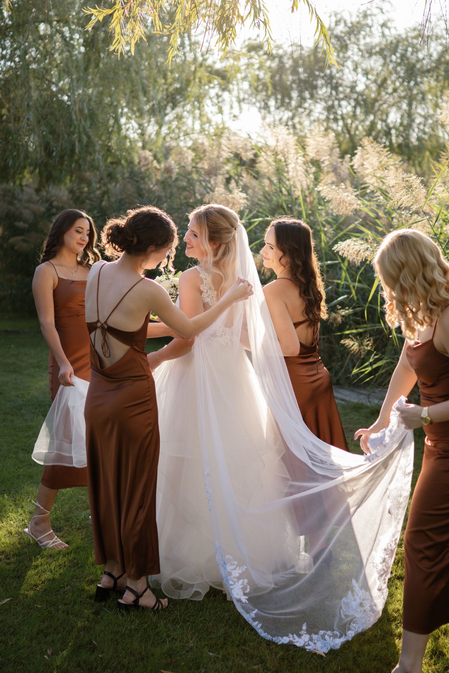 Wedding party in brown bridesmaid dresses helping with white bridal gown outdoors.