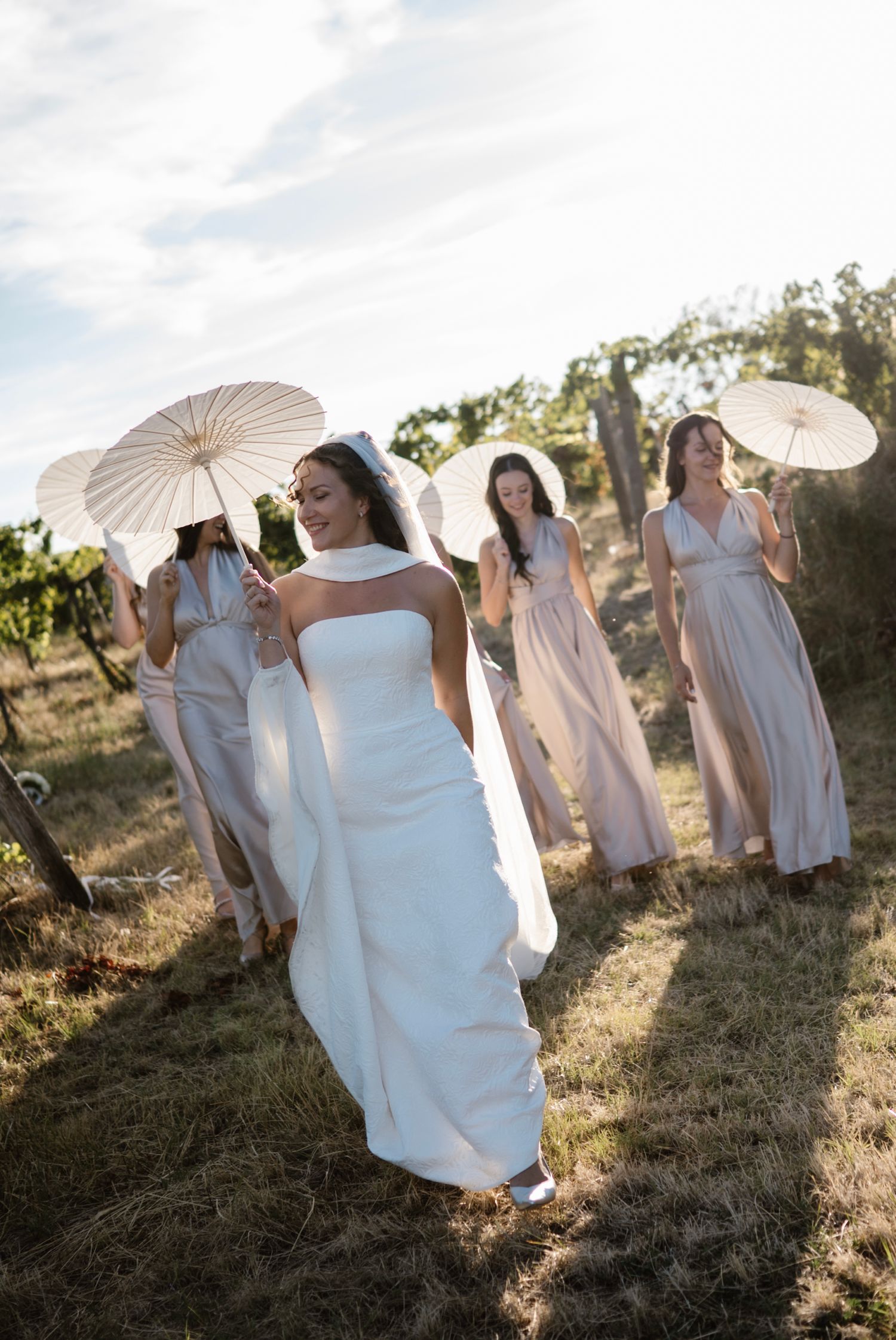 Bride in white gown leads bridesmaids in blush dresses holding parasols through a sunlit vineyard.