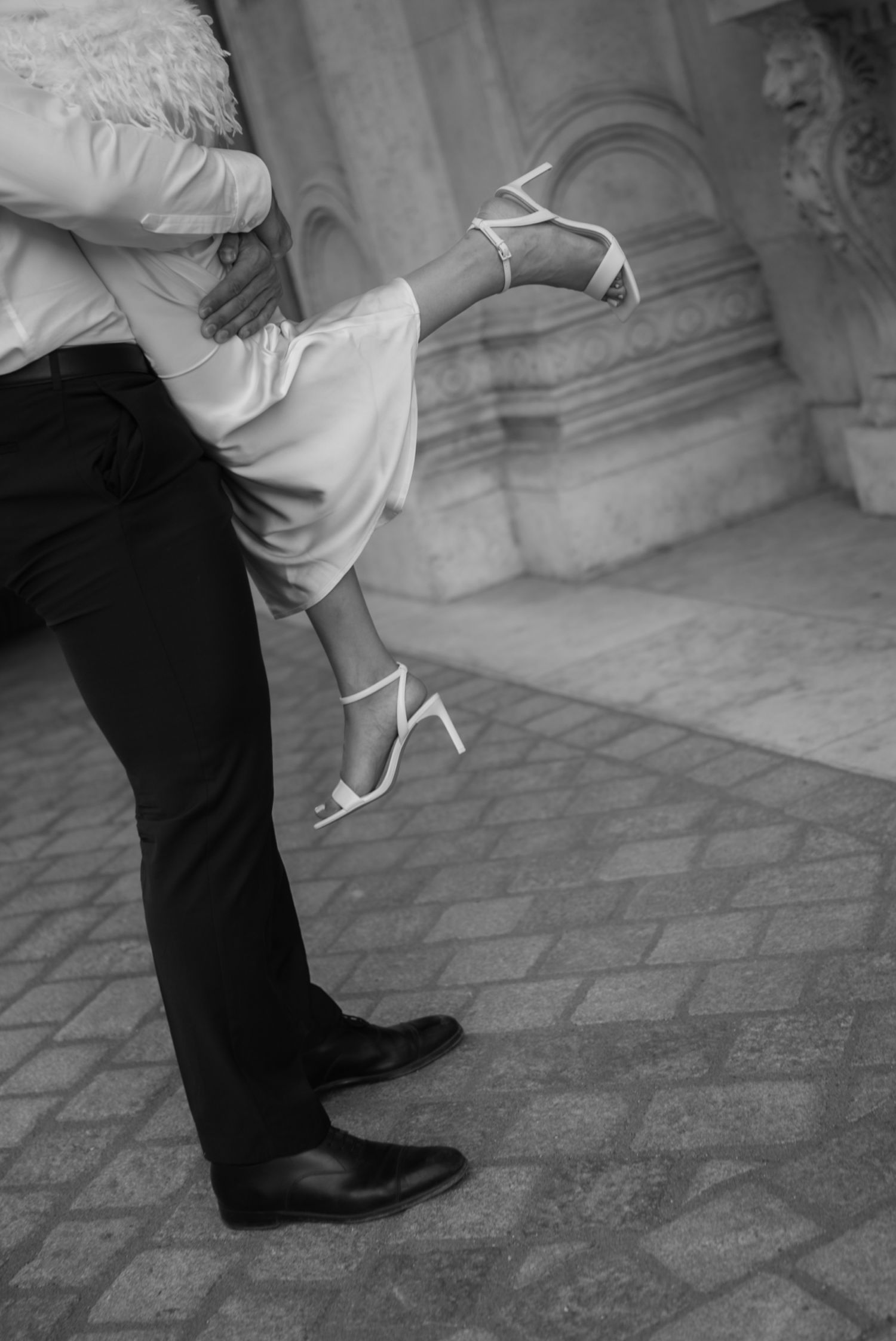 Black and white photo of a couple embracing, bride in white heels lifted by groom in dark suit on stone pavement.