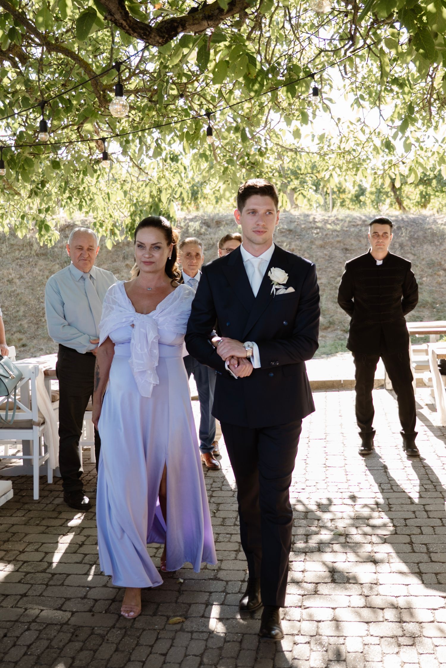 Couple walking down outdoor wedding aisle under green tree canopy.