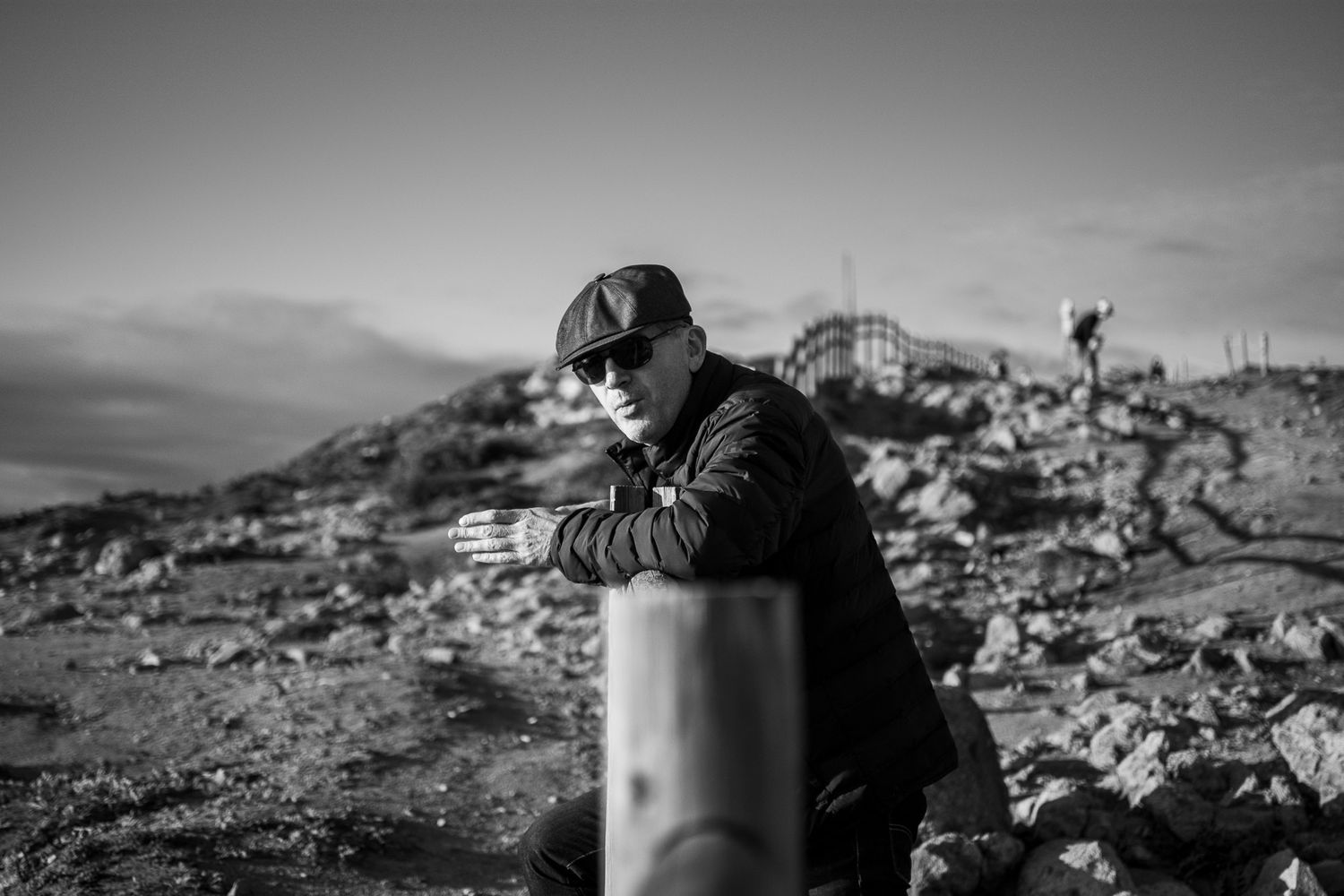 A black and white photo of a figure leaning on a metal post against a rugged mountain backdrop.
