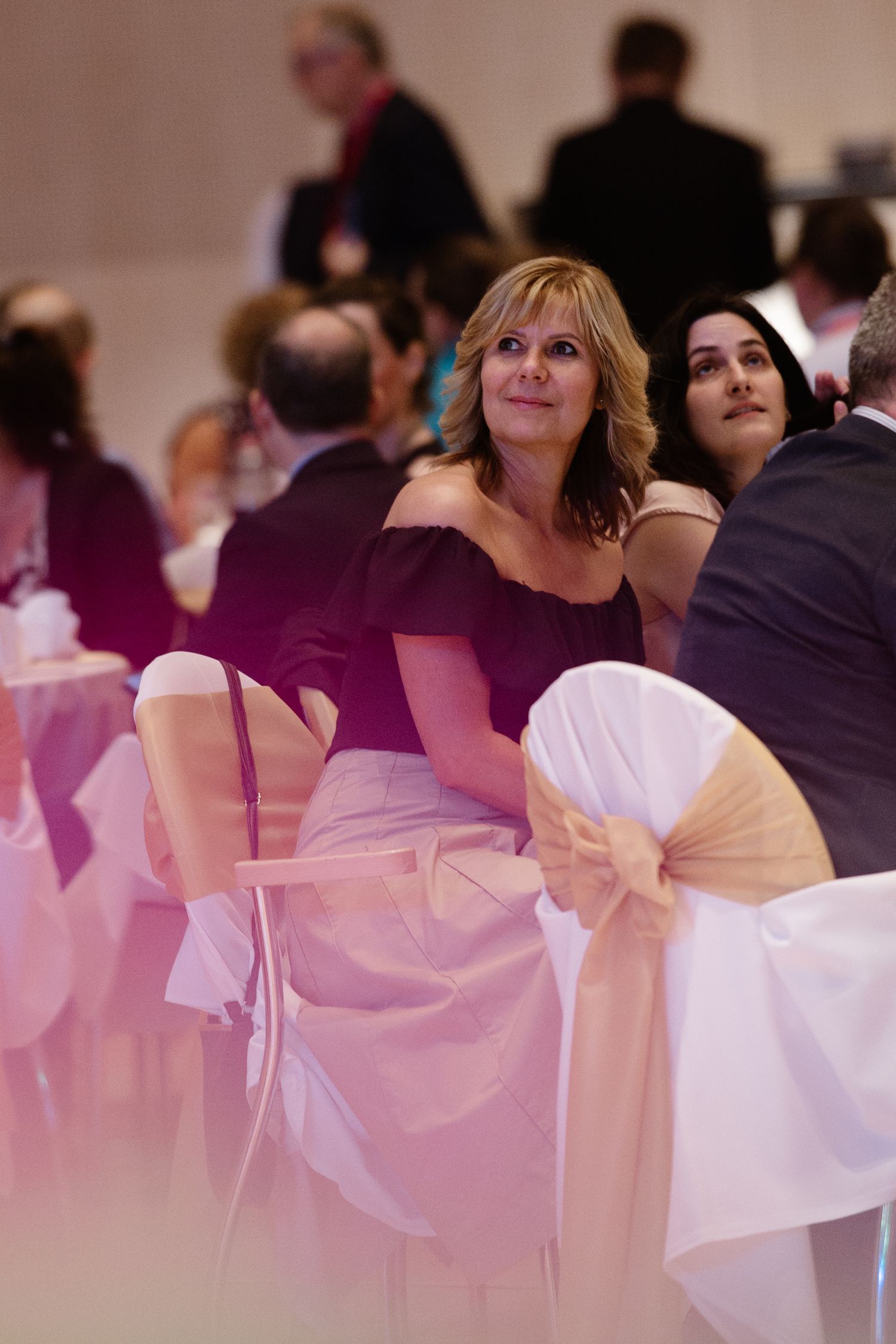 Guests seated at formal event with pink chair decorations and white bows in an elegant banquet setting.