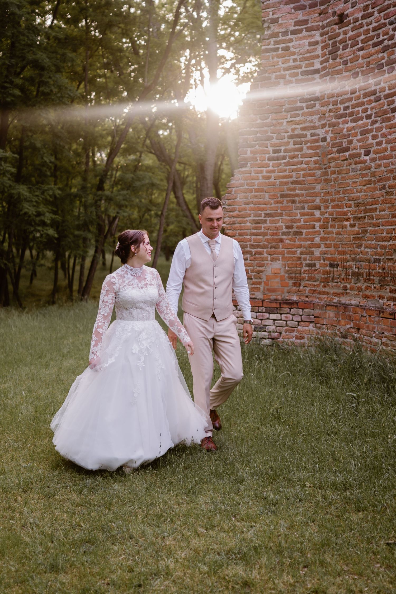 Newlyweds walk hand in hand along brick wall during golden hour wedding photoshoot.
