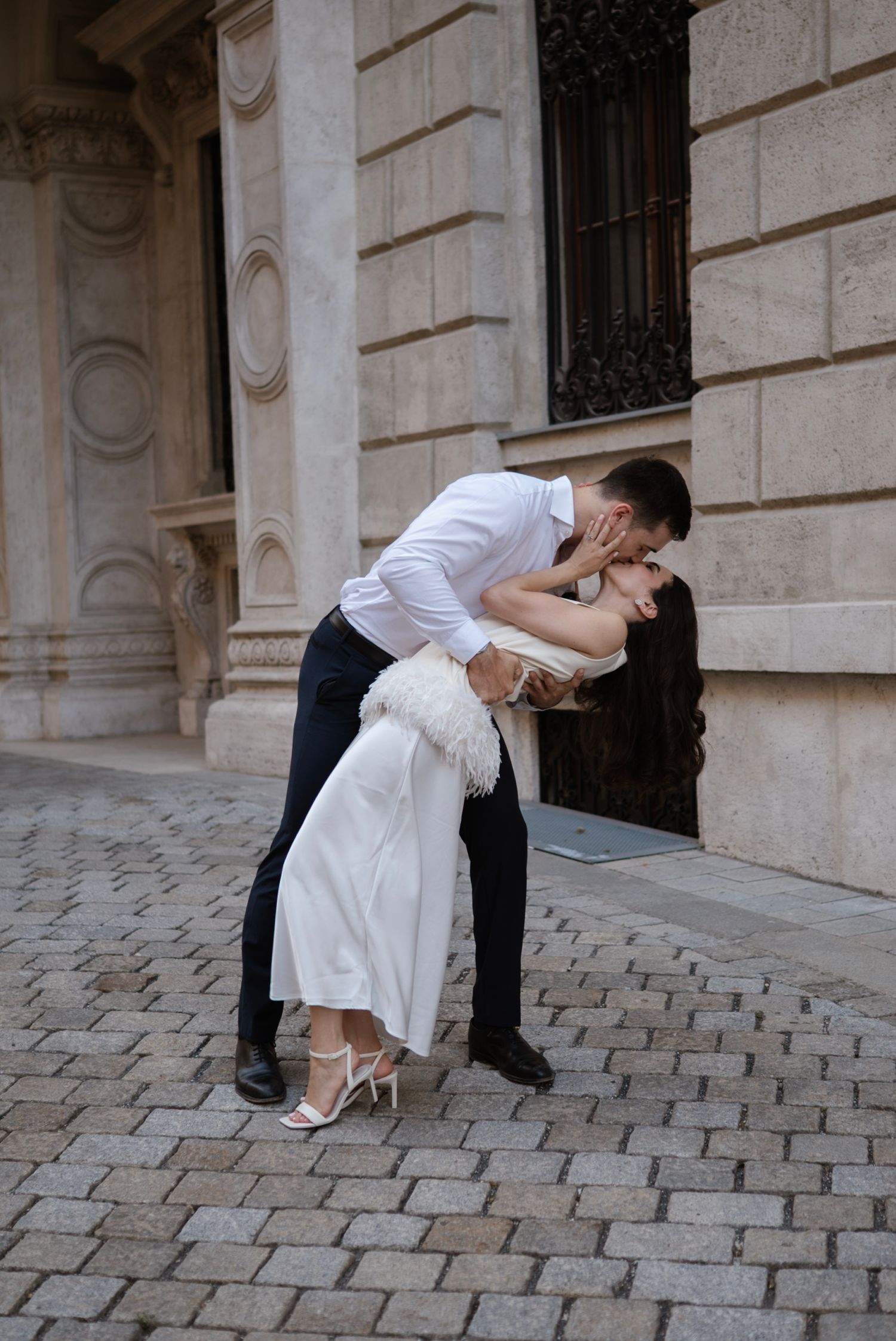 A couple shares a romantic dip kiss on cobblestone street, bride in white dress and groom in white shirt.