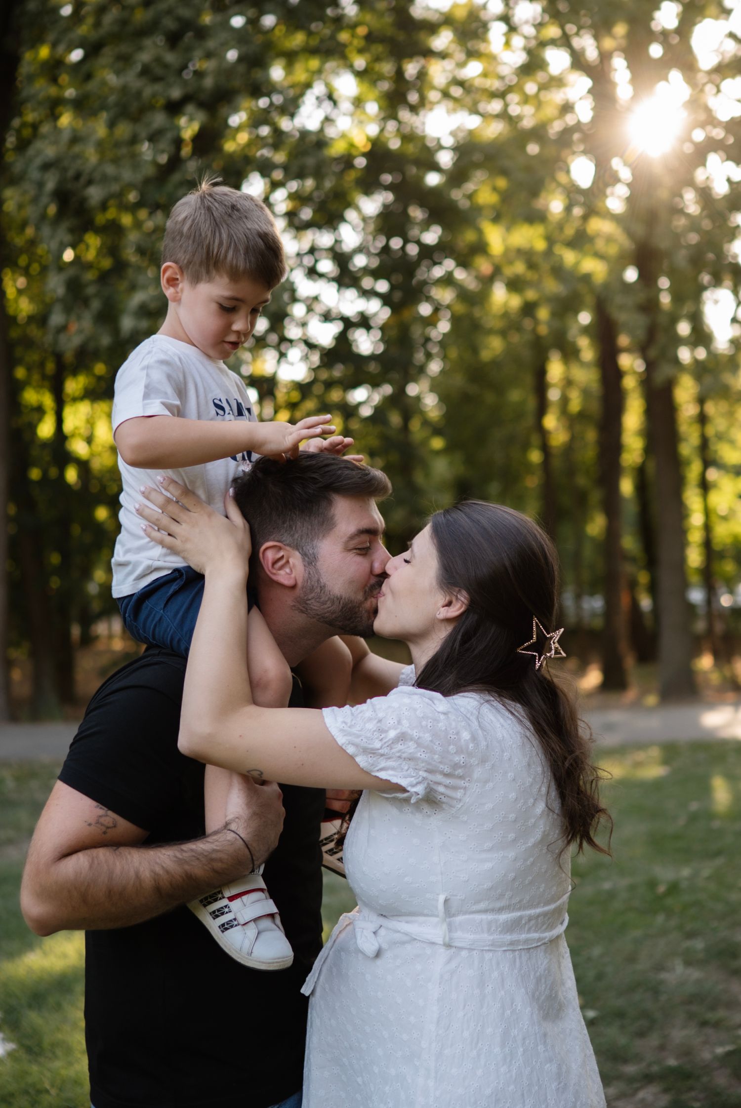 A couple kisses in a sunny park while their young son sits on the mans shoulders, captured in warm golden light.