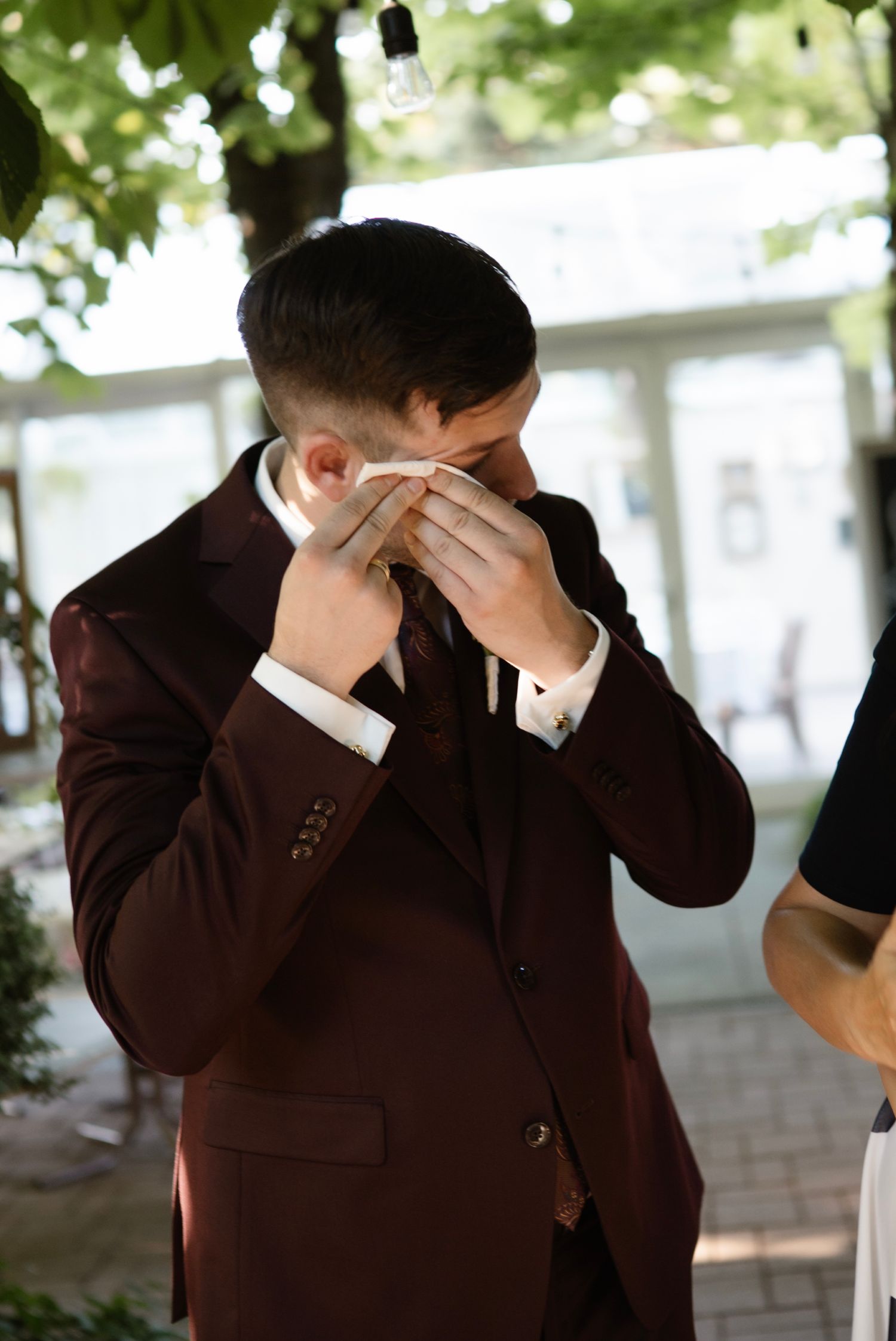Emotional groom in burgundy suit wiping tears with handkerchief at wedding ceremony.