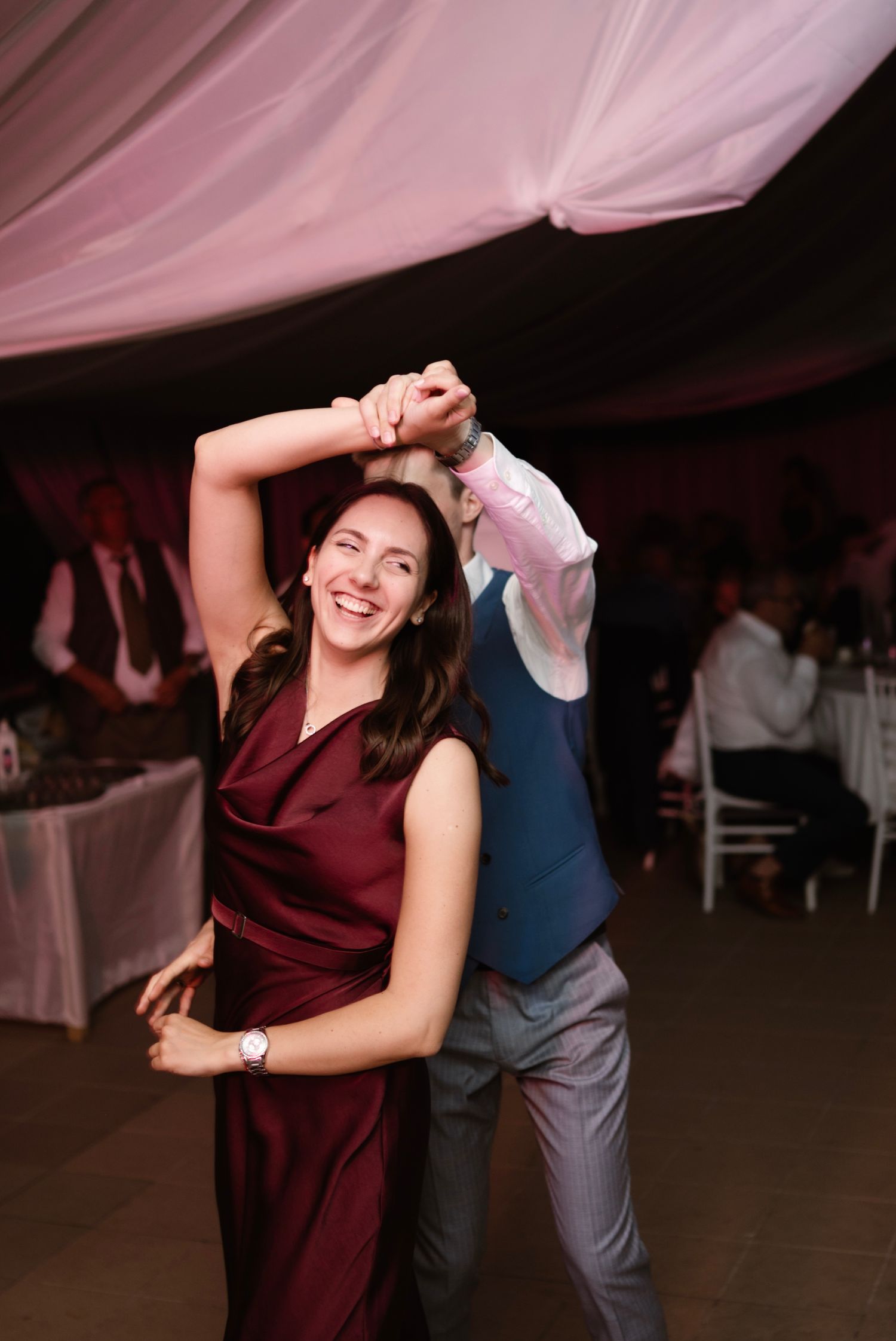 A woman in a burgundy dress laughs while dancing with a partner at an elegant wedding reception under pink draped ceilings.