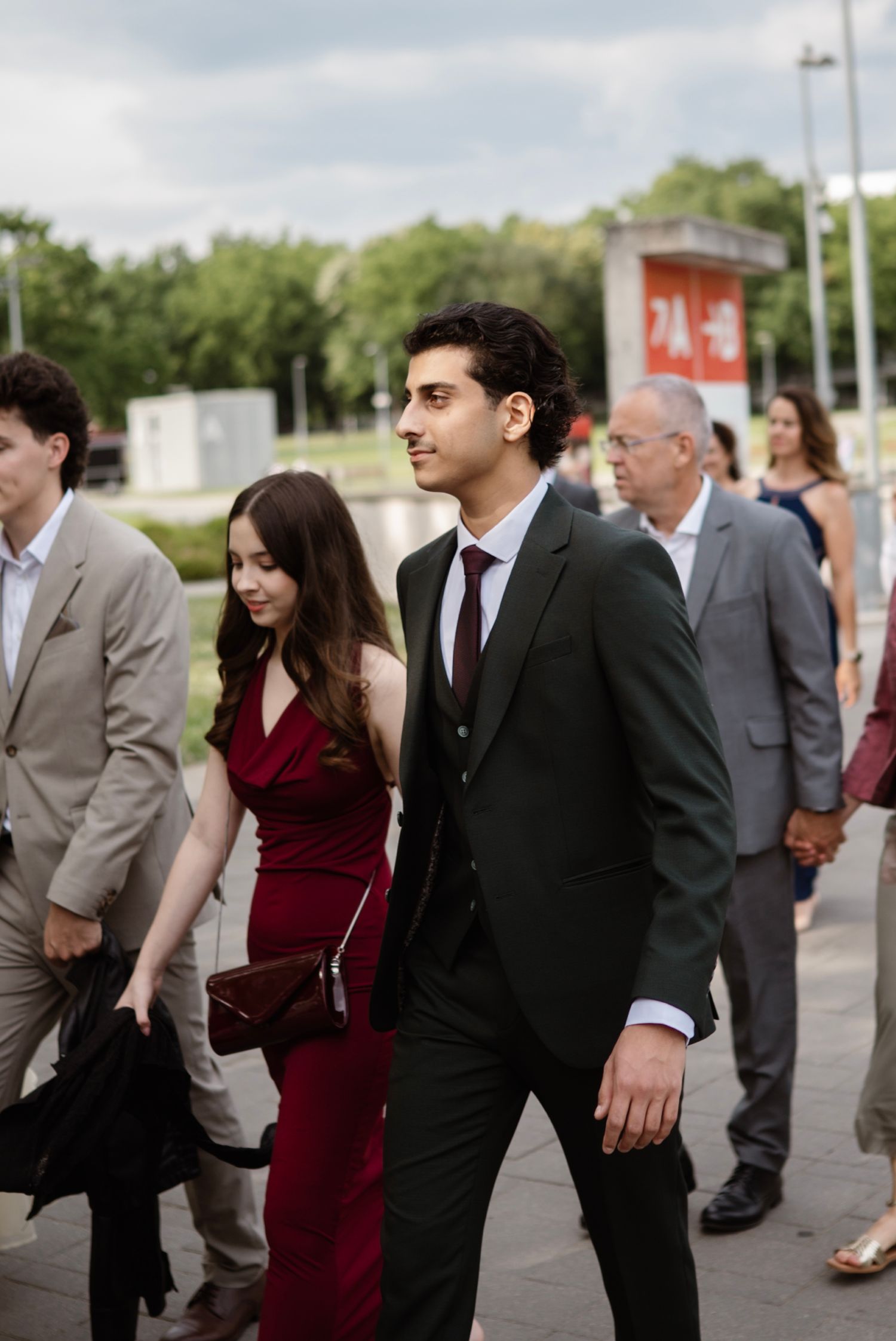 Elegant couple in formal attire walk together outdoors, man in dark suit with burgundy tie, woman in red dress.