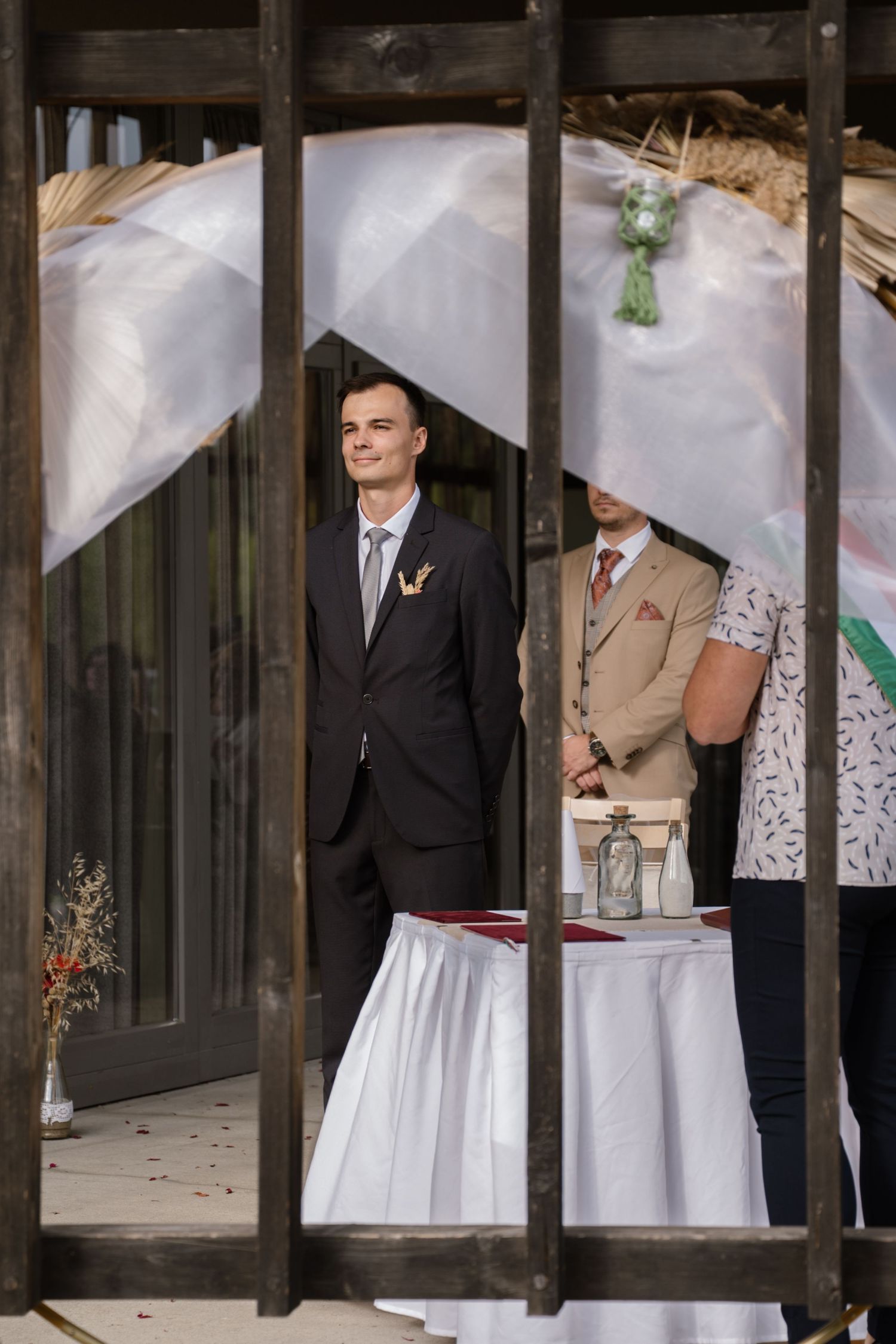 Wedding ceremony setup viewed through wooden beams with attendees in formal attire.