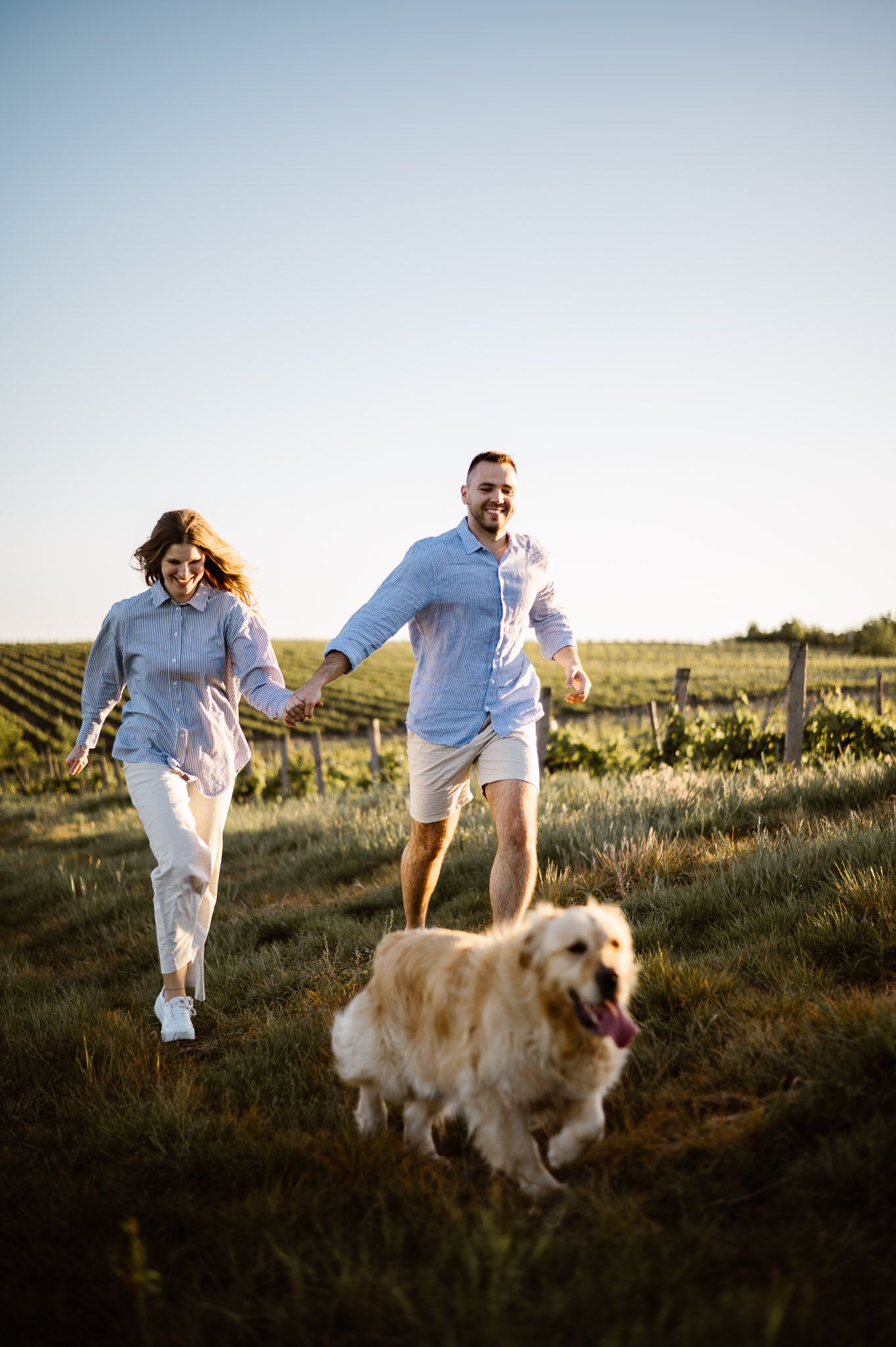 Playful couple running with their golden retriever through a scenic field at sunset.
