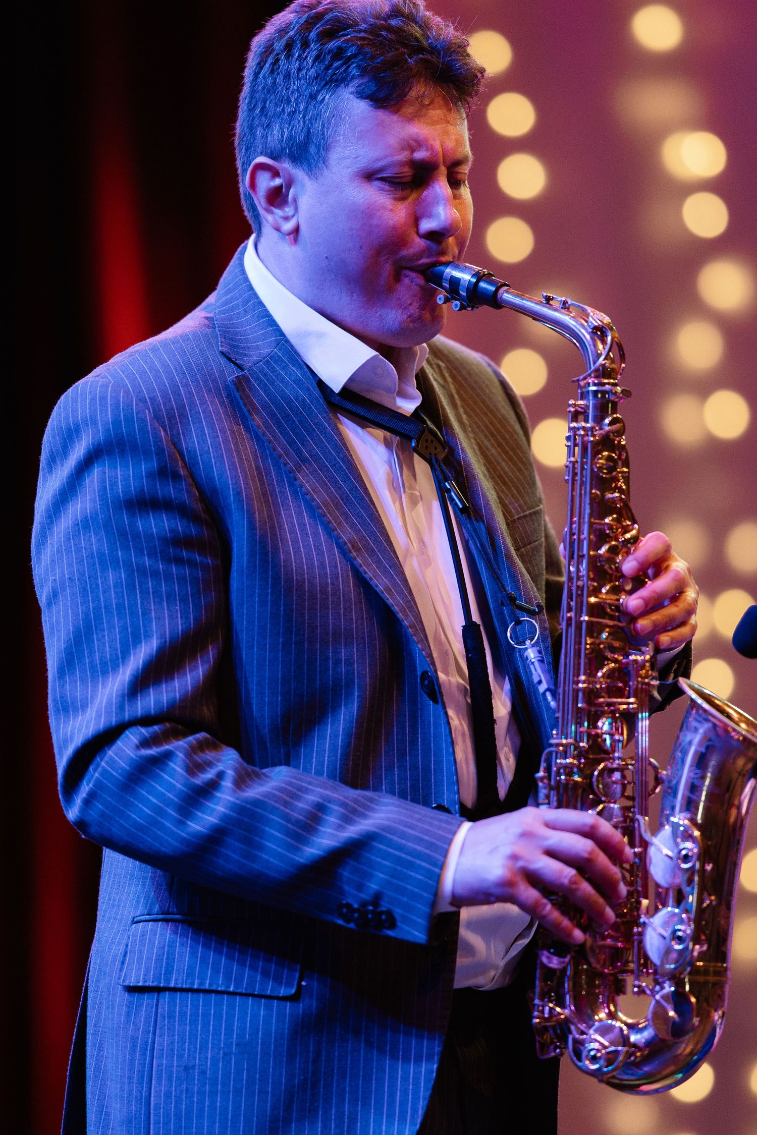 Saxophonist in pinstripe suit performing against backdrop of blurred stage lights.
