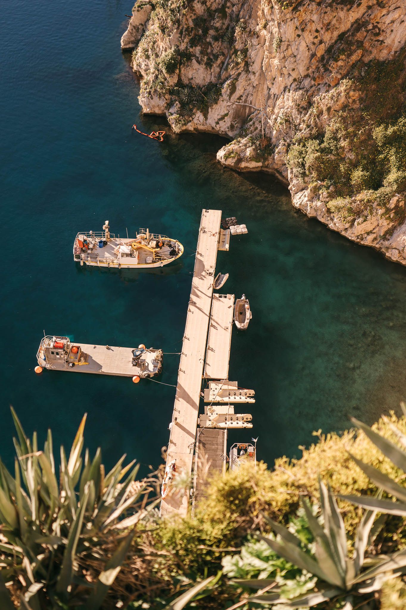 Aerial view of a diving platform and dock extending into crystal blue Mediterranean waters near rocky cliffs.