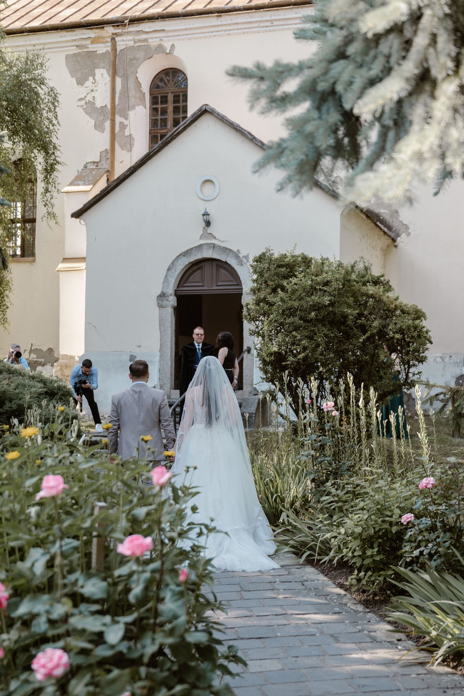 A bride in white walking towards a charming chapel entrance surrounded by garden flowers.