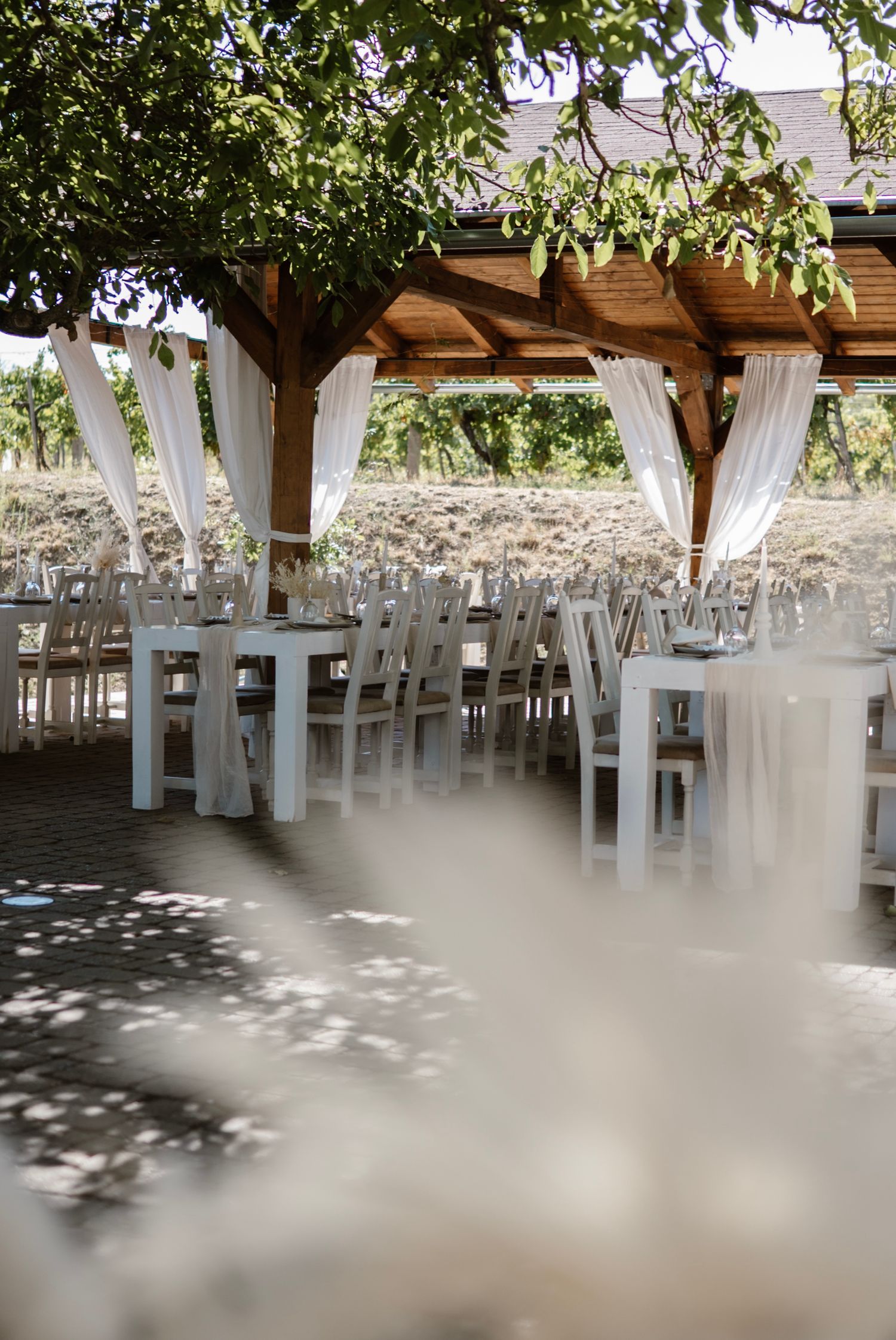 Elegant outdoor wedding reception setup with white chairs and tables under a wooden pergola with flowing white drapes.