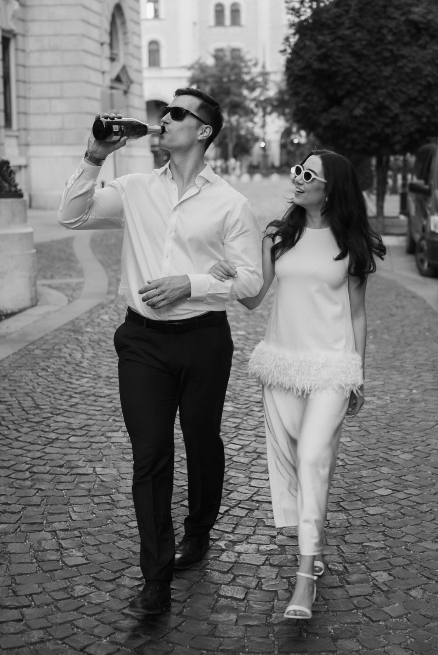 Stylish couple walking on cobblestone street, man drinking from bottle, woman in white dress, black and white photo.