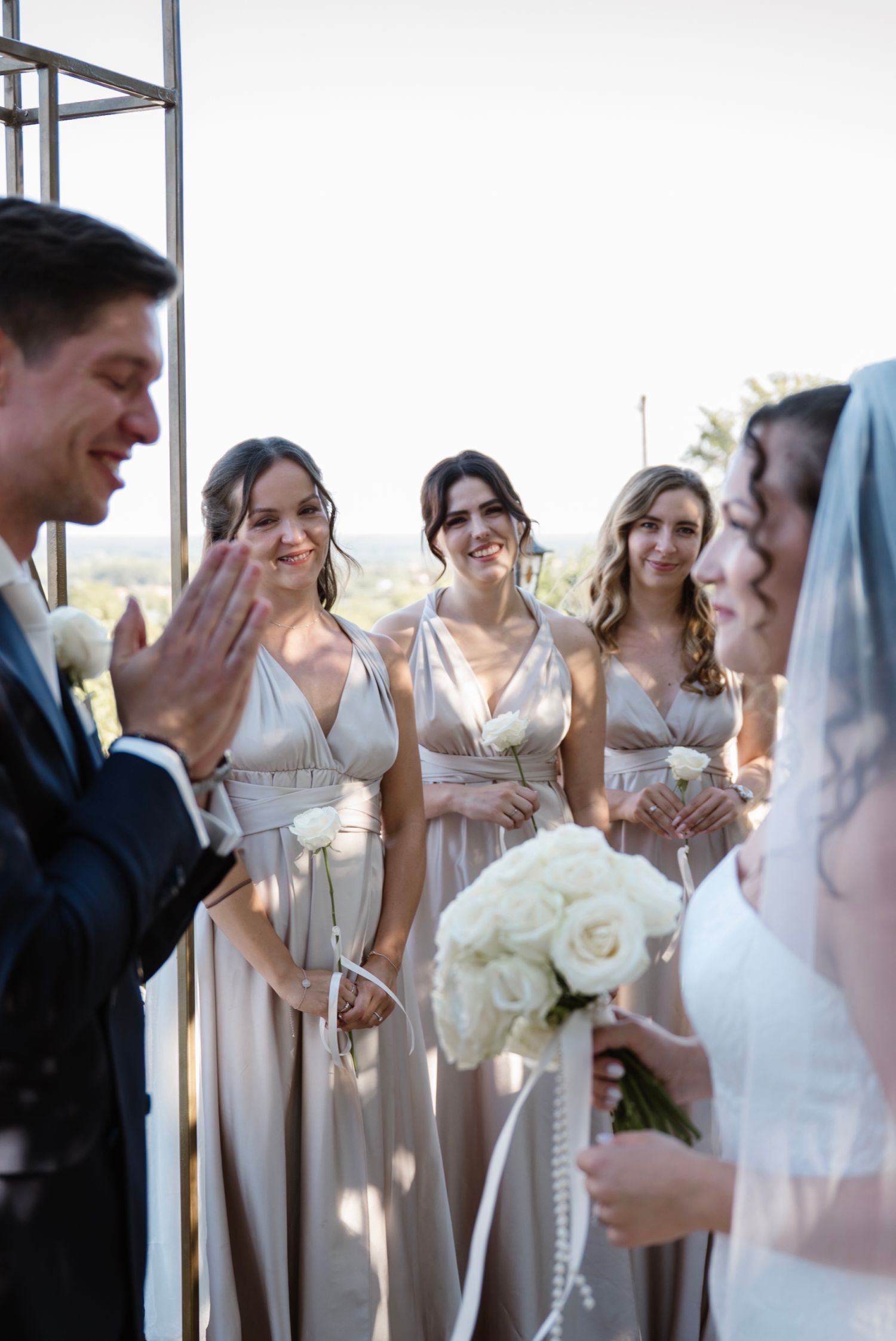 Bride holding white rose bouquet faces groom as smiling bridesmaids in champagne gowns look on at outdoor wedding ceremony.