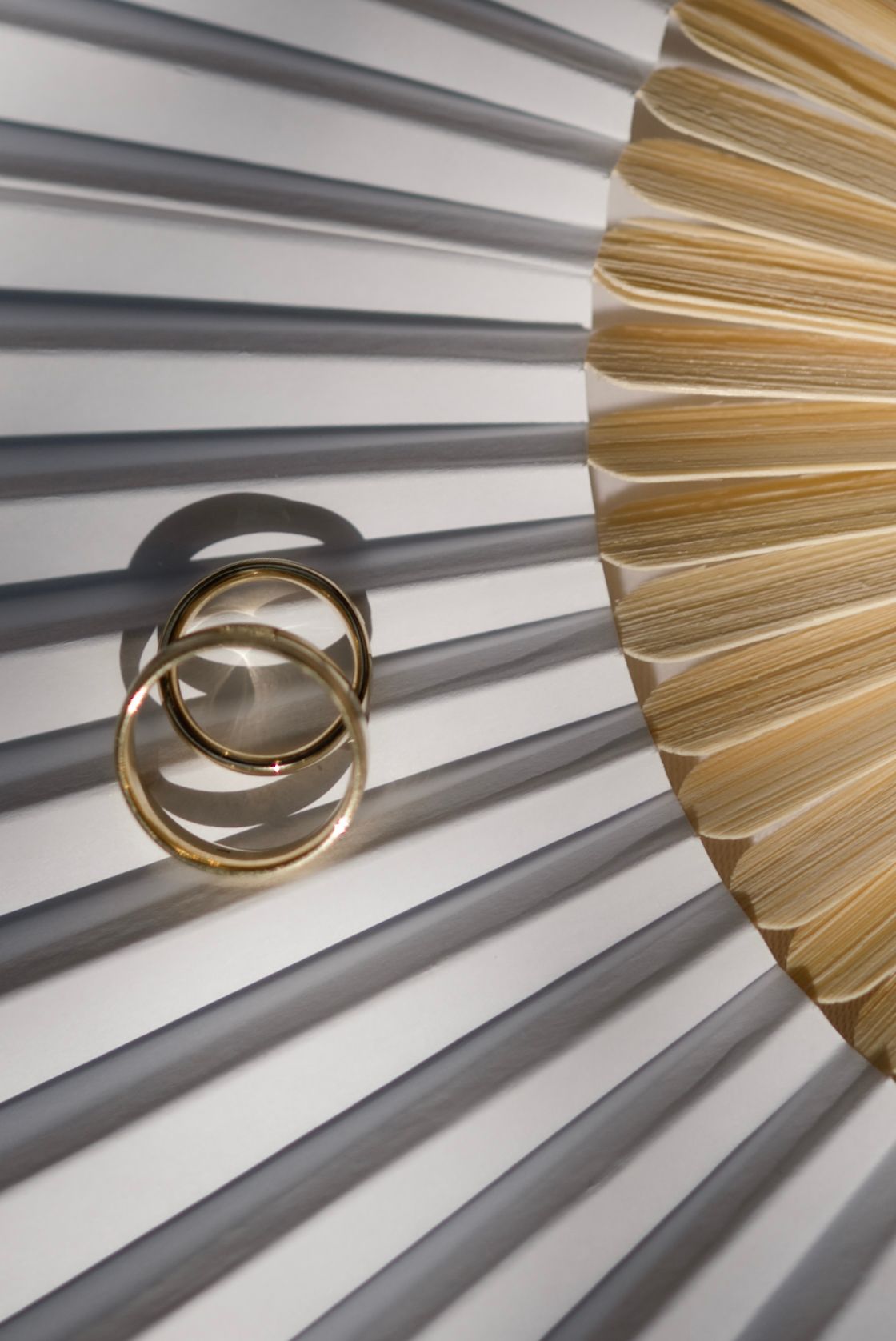 Two gold wedding bands resting on pleated white fabric beside a golden fan-shaped decorative element.