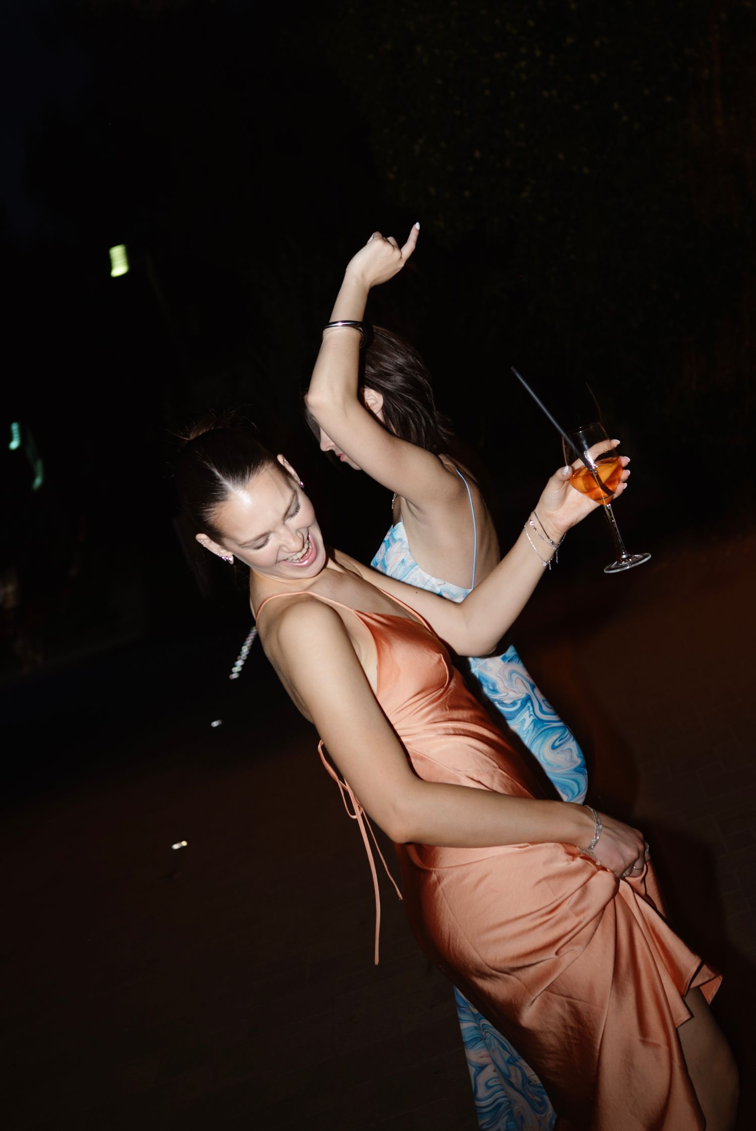 Young woman in elegant peach satin dress dancing and holding a cocktail glass at an outdoor evening party.