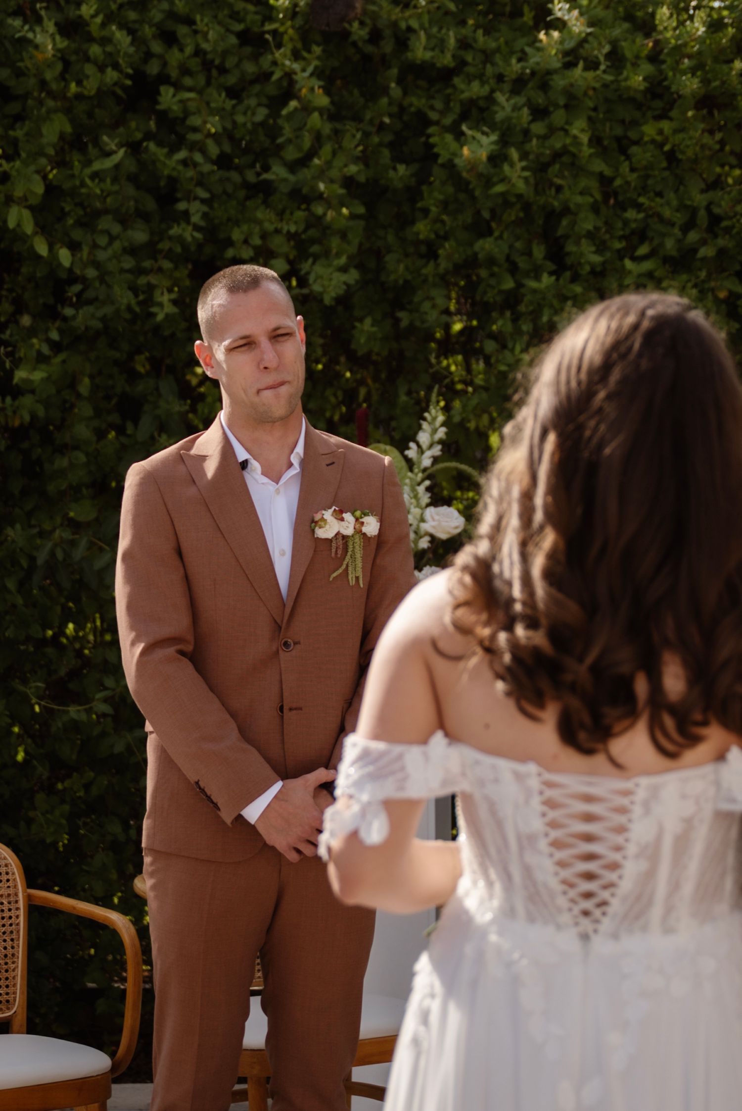 Emotional groom in brown suit watches his bride approach during an outdoor wedding ceremony.