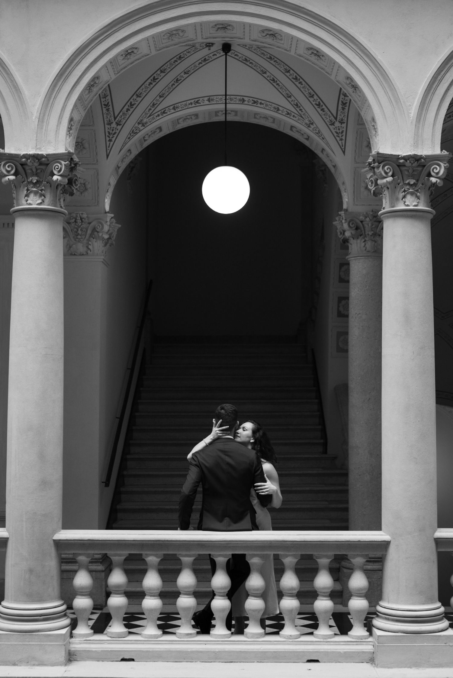 Couple embracing on grand staircase under glowing orb light, framed by ornate arches and columns, black and white photo.
