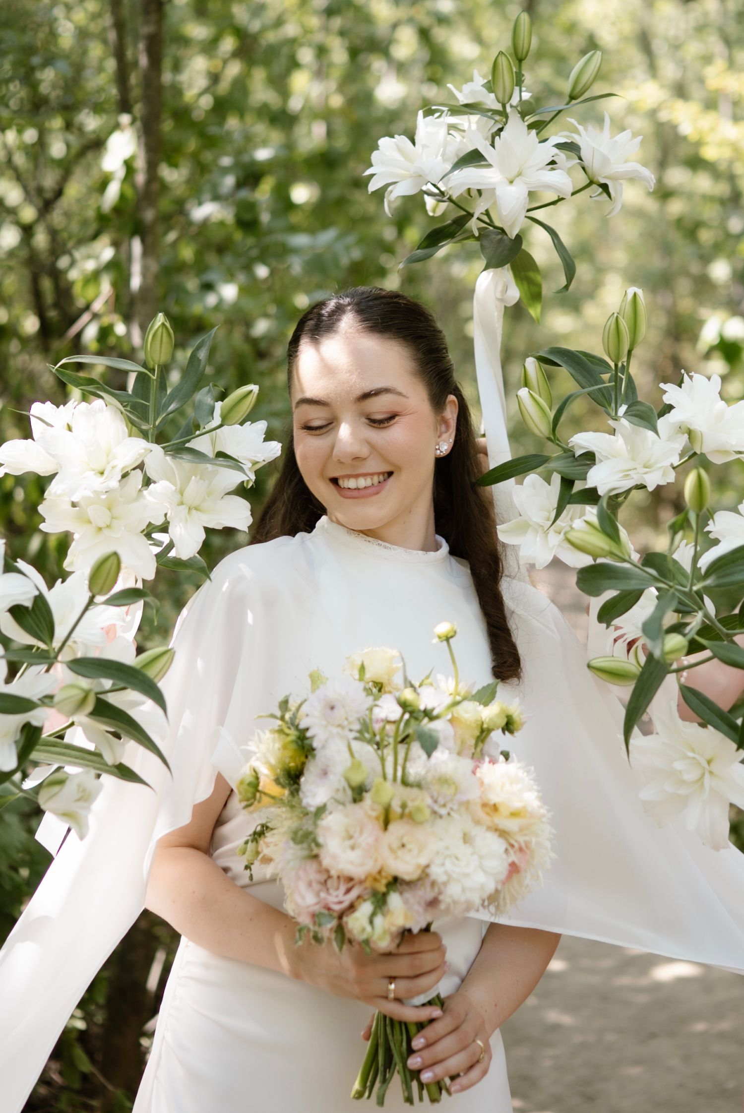 Smiling bride in white dress holding pastel bouquet surrounded by white lily flowers in a lush green garden setting.