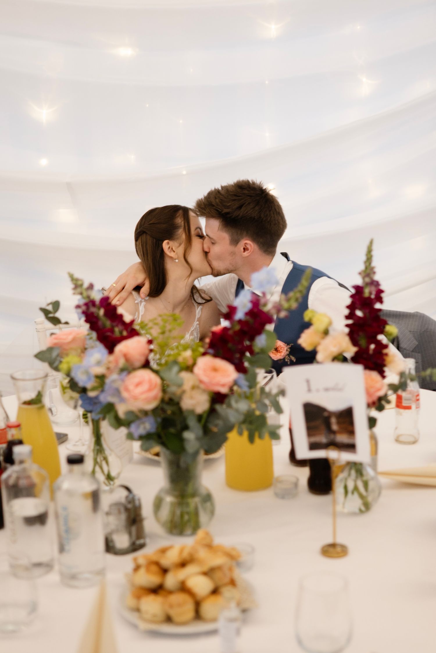 Newlyweds share a romantic kiss at their wedding reception table adorned with colorful floral arrangements.