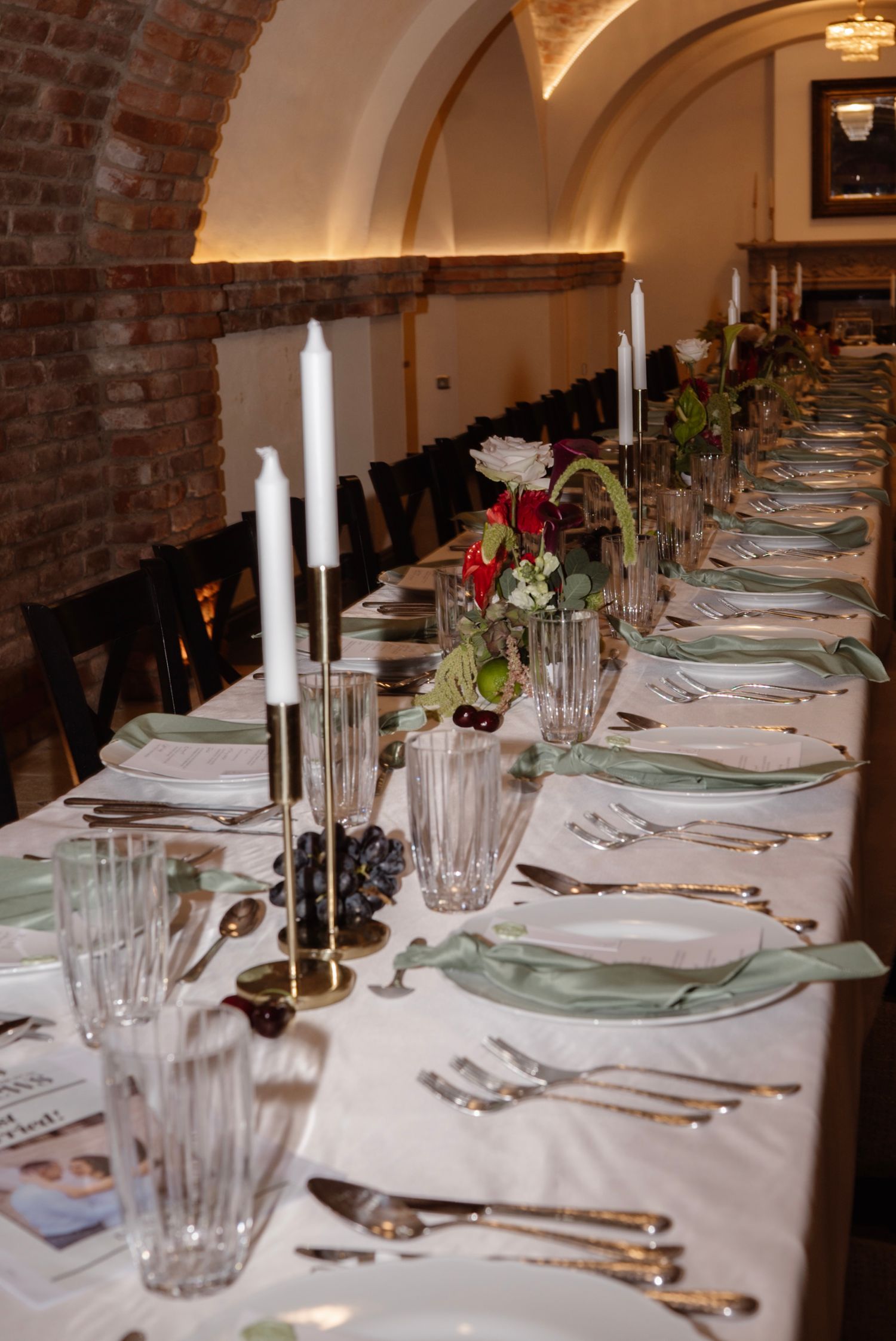 Elegant long dining table set with white candles, crystal glasses, green napkins, and floral centerpieces in a brick-arched venue.