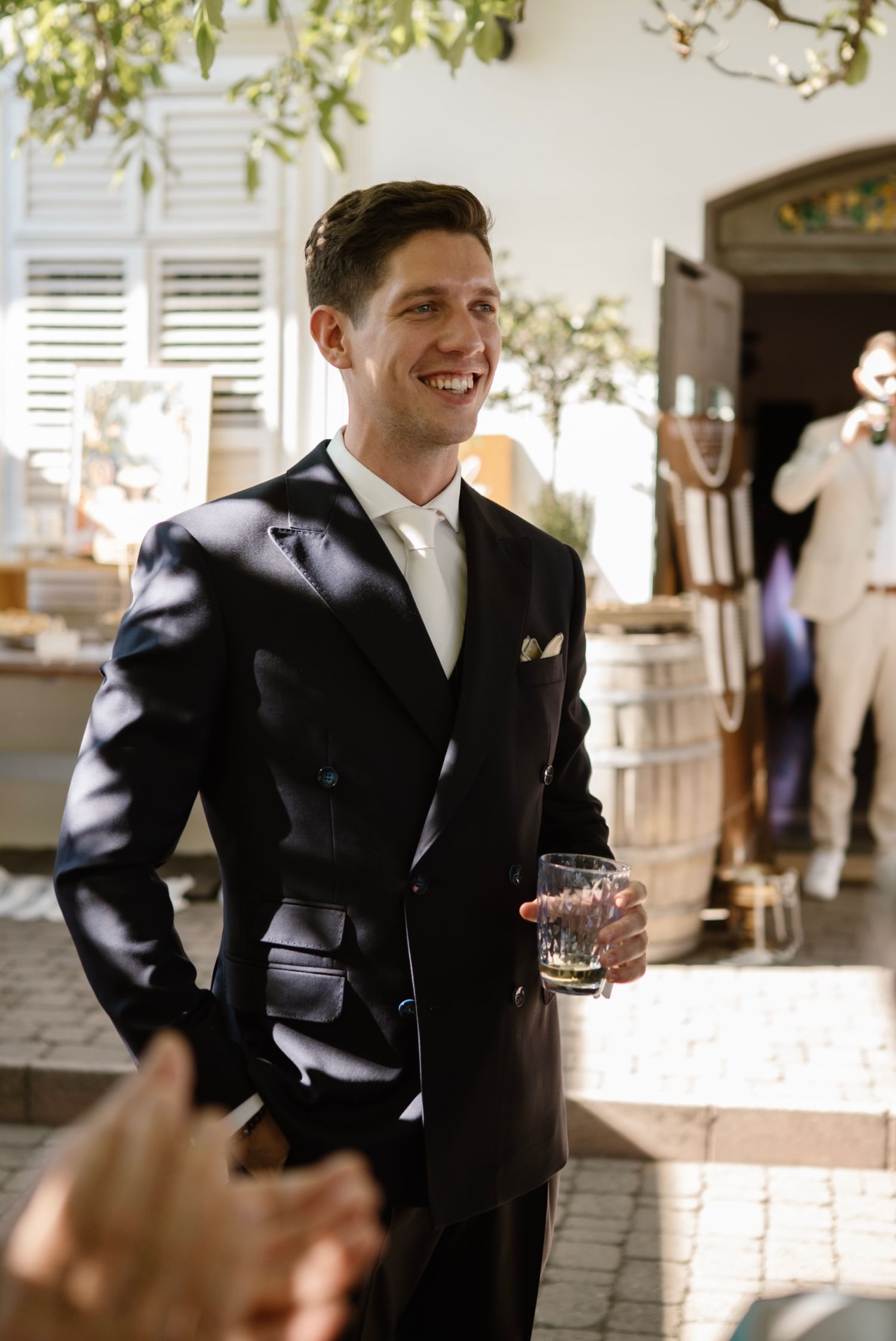 Smiling groom in a dark suit holding a drink outdoors at a sunny wedding reception venue.