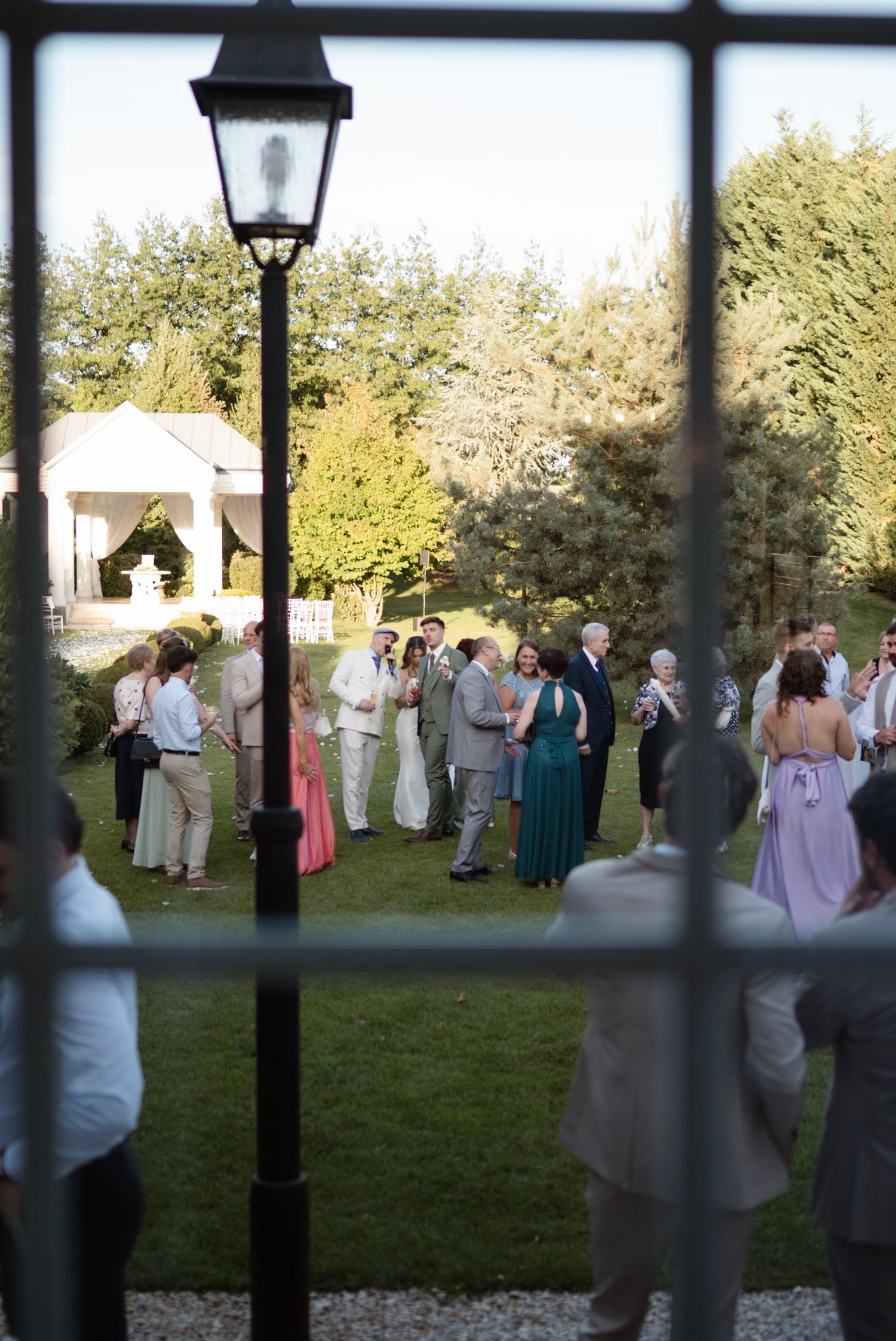 Wedding guests gather outdoors on a lawn, viewed through a window, with a lamp post and lush greenery in the background.