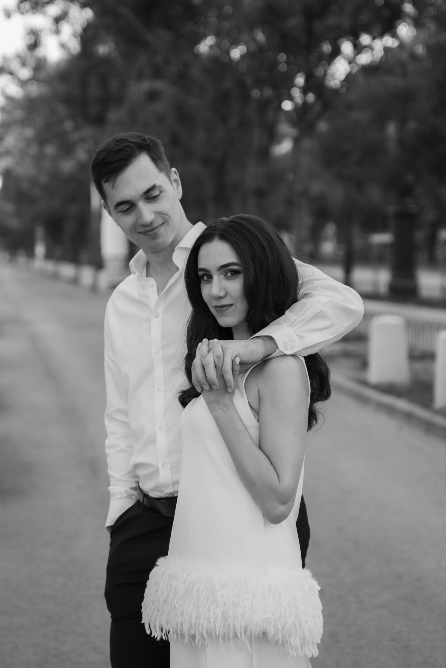 Black and white photo of a romantic couple posing outdoors, man in white shirt embracing woman in fringe dress.