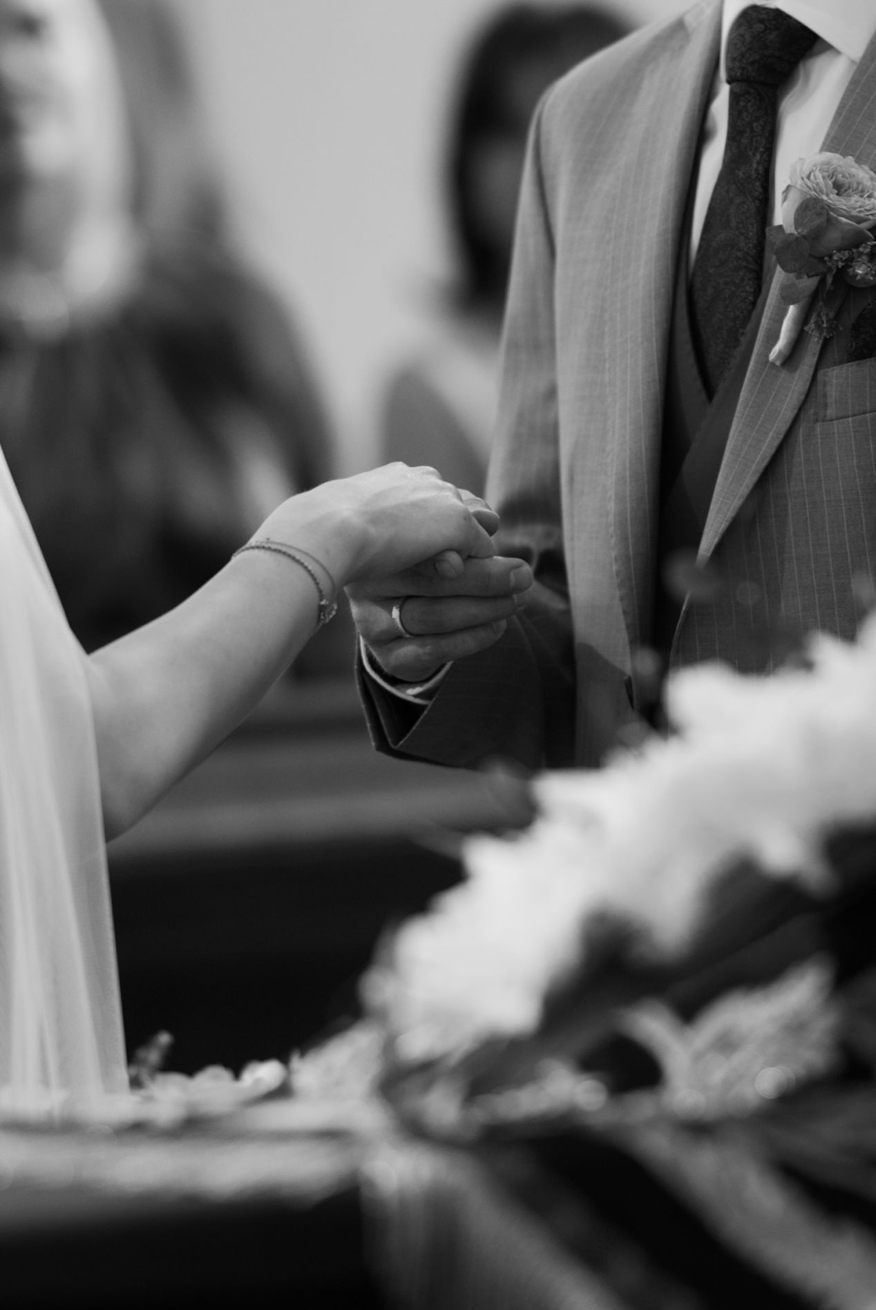 Black and white wedding photo of bride and groom holding hands at the altar with flowers in the foreground.
