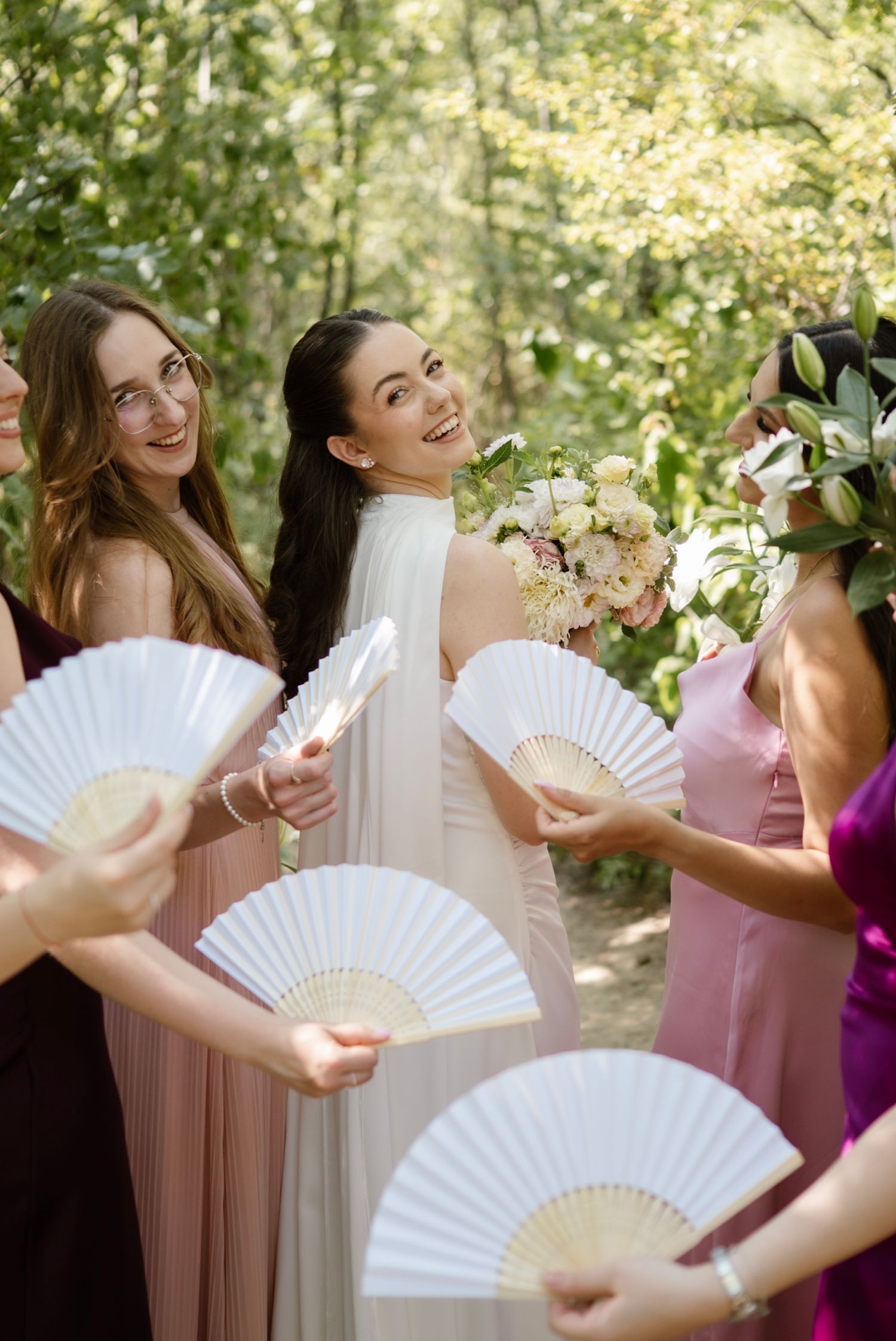 Bride holding floral bouquet smiles with bridesmaids in pink dresses holding white hand fans outdoors.