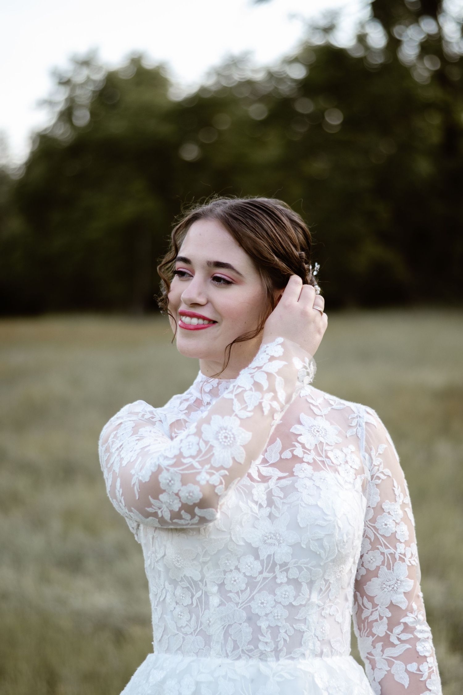 Bride in lace wedding dress smiles joyfully while adjusting her hair outdoors.