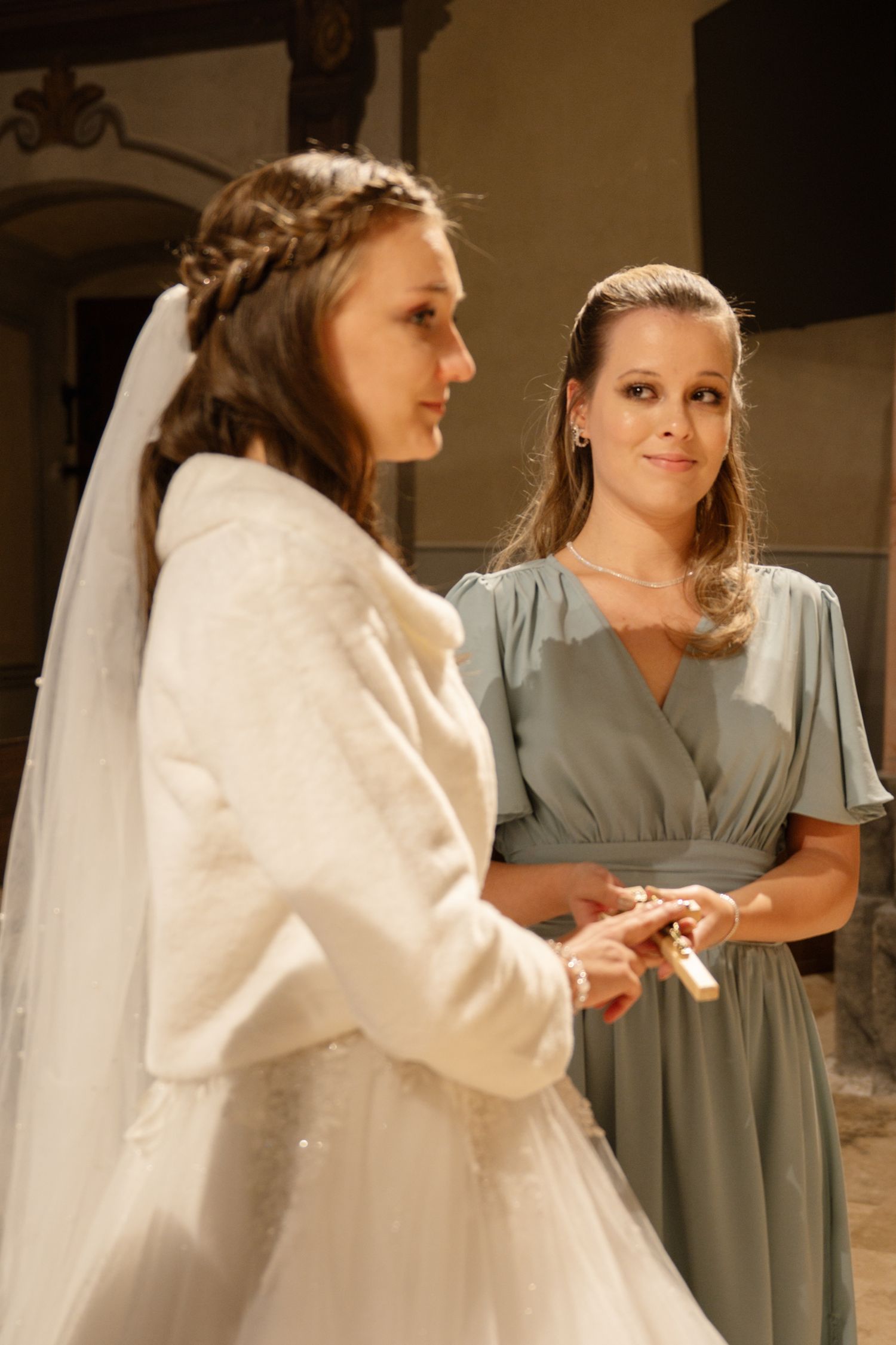 A bride in a white dress and veil stands with an attendant wearing a sage green dress.