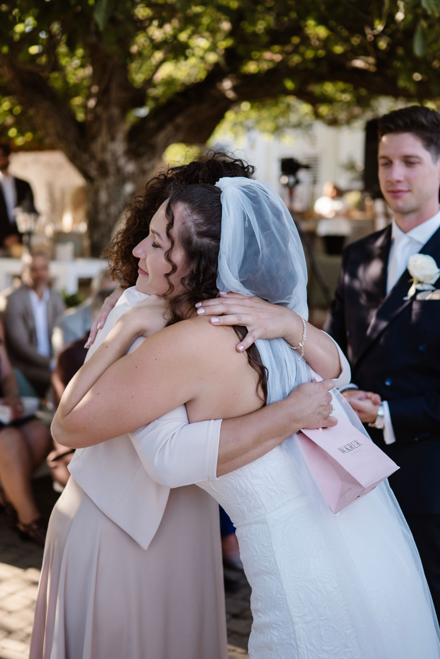 Bride in white gown and veil sharing emotional hug with bridesmaid during wedding ceremony.