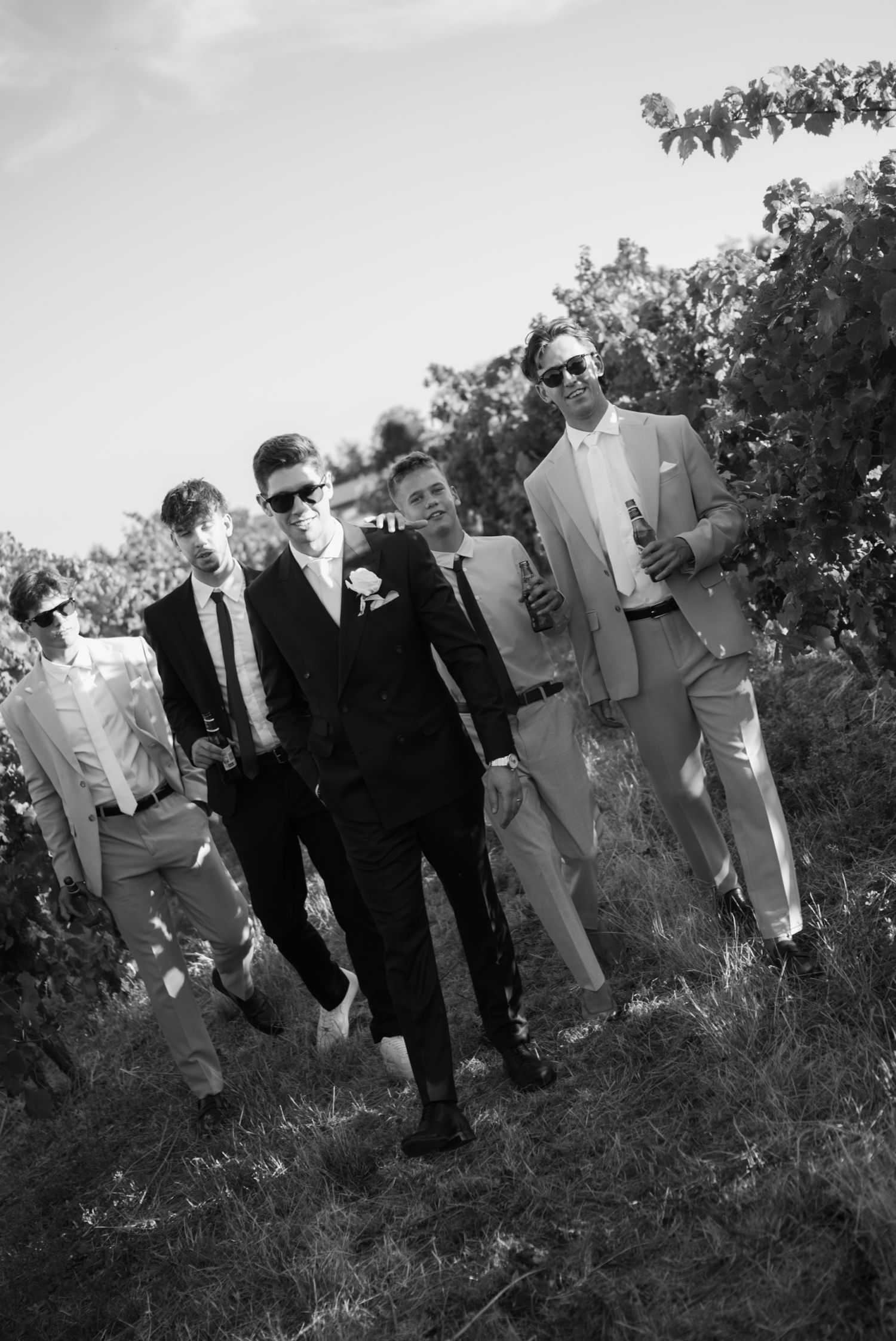 Groom and groomsmen in suits walk through vineyard in black and white wedding photo.