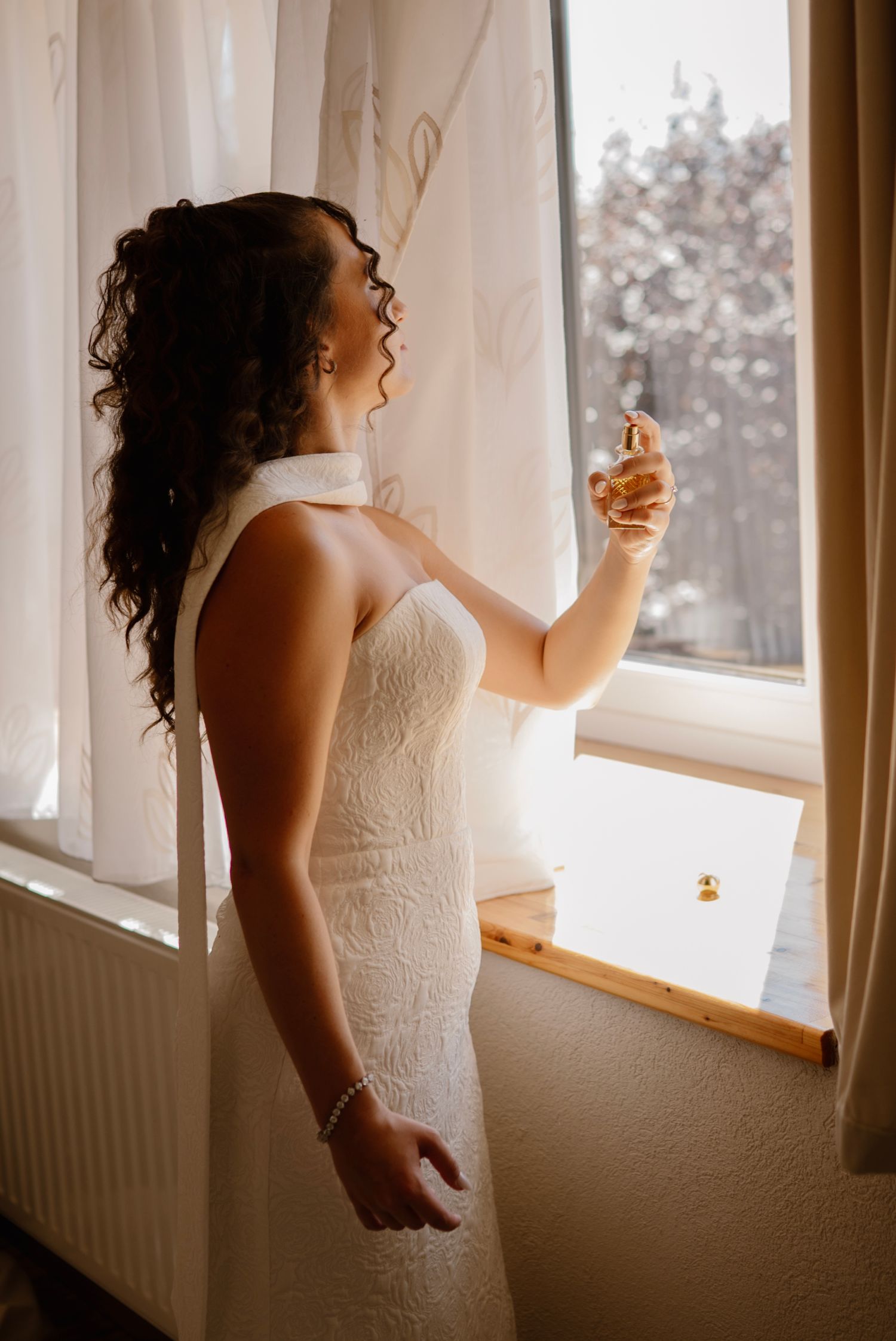 Bride in white lace gown spraying perfume by a sunlit window on her wedding day.