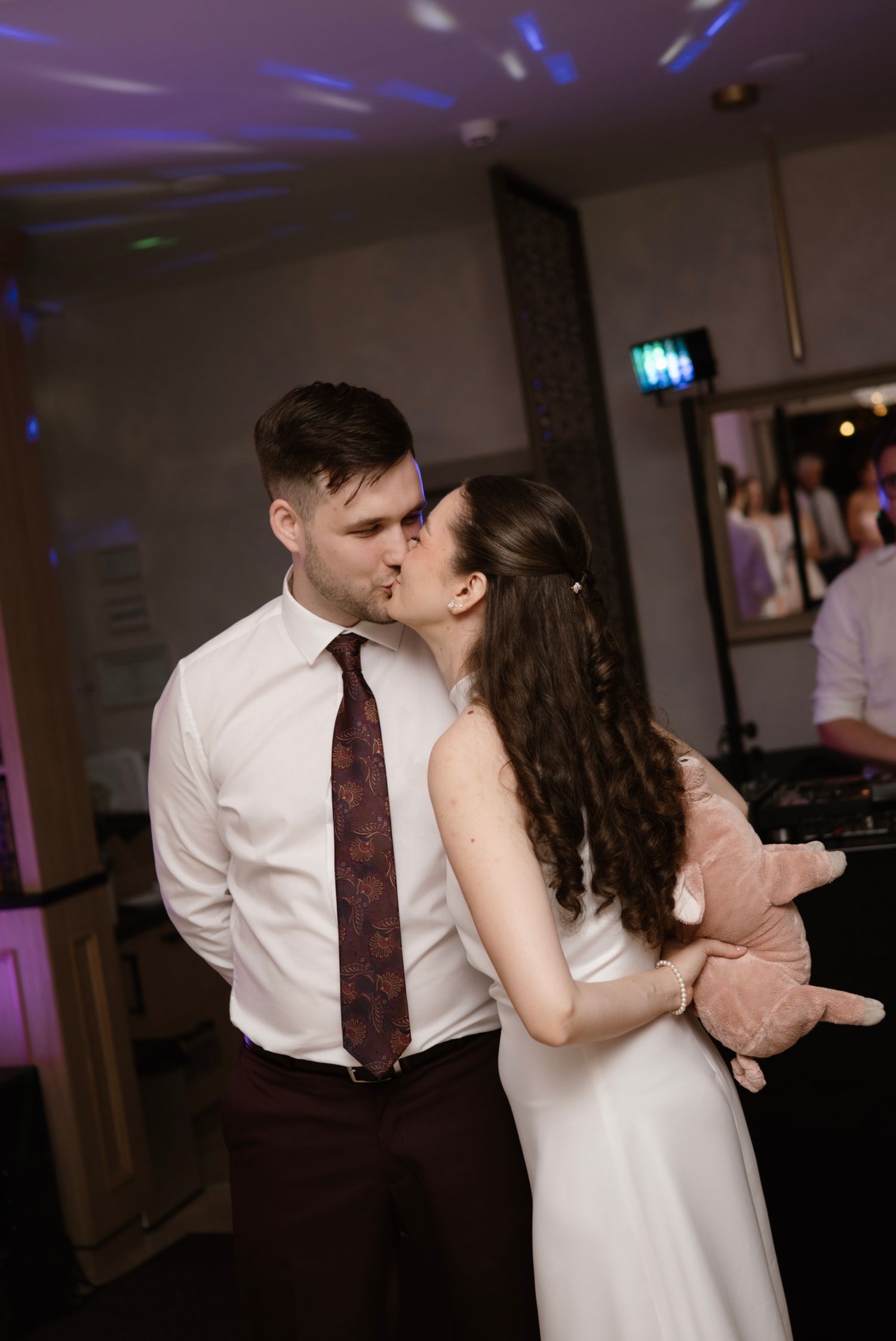 Bride and groom sharing kiss on dance floor while bride holds stuffed animal.