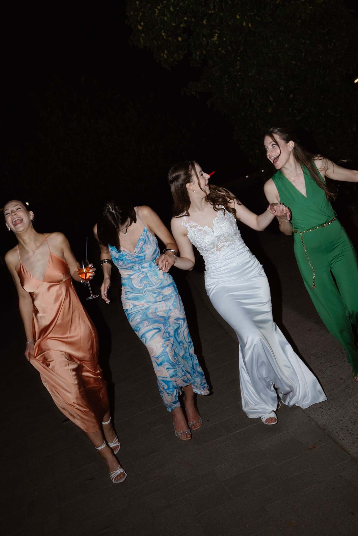 Four women in elegant formal dresses dancing joyfully together at an outdoor evening event.