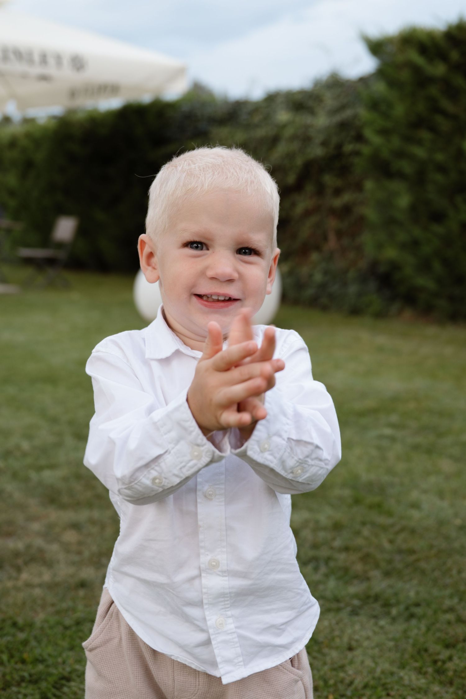 Small child in white dress shirt claps and smiles on grassy lawn.