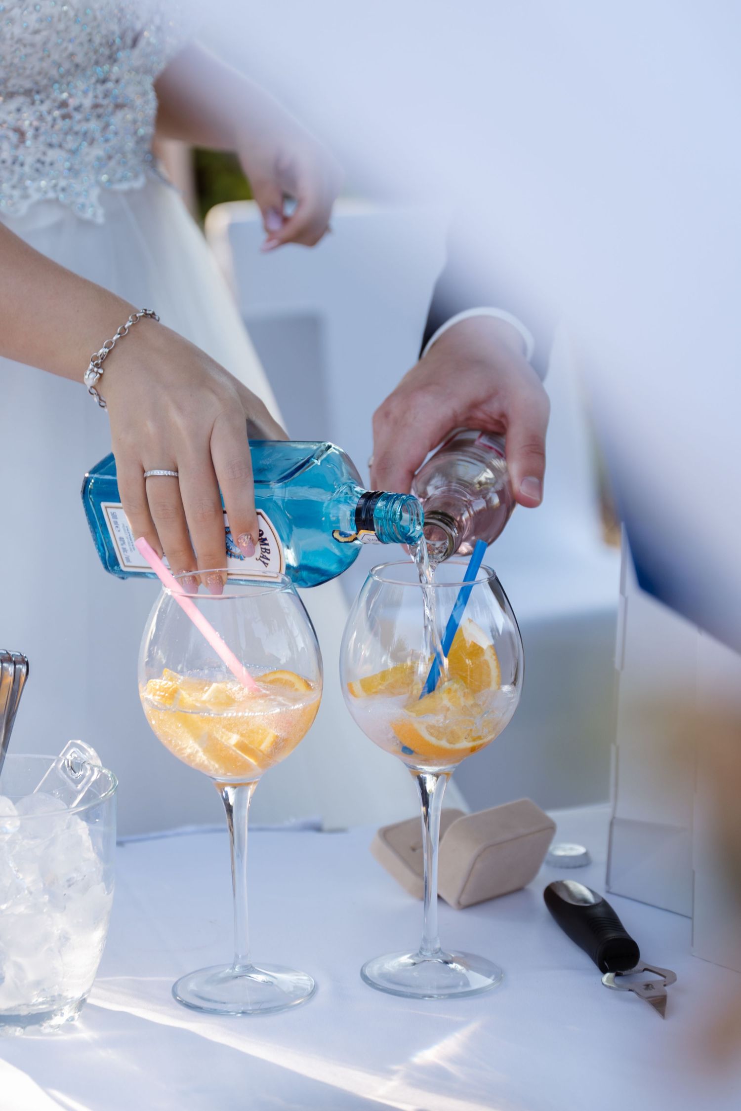 Hands pour blue cocktails into elegant wine glasses on a white surface.