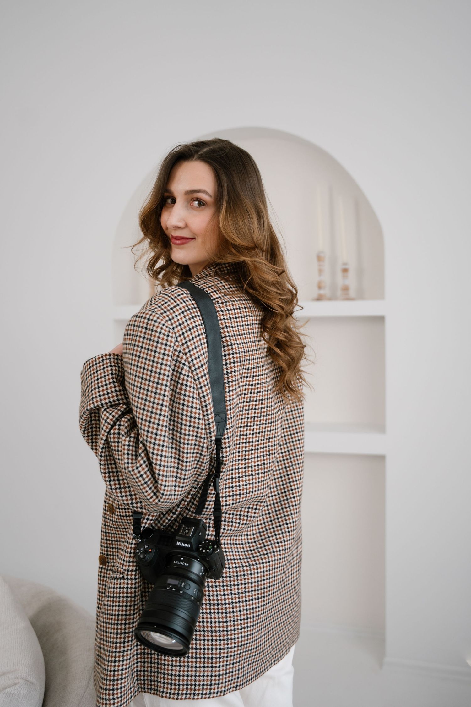 A person wearing a brown checkered blazer poses with a professional camera against a white minimalist background.