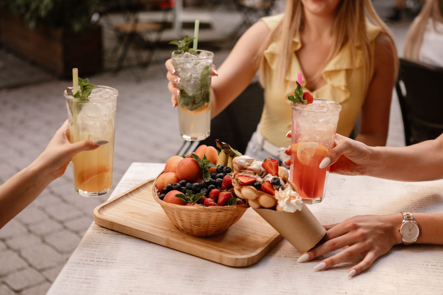 Friends enjoying craft cocktails and appetizers at an outdoor restaurant patio on a sunny afternoon.