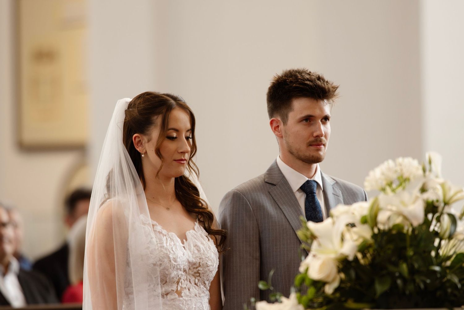 Bride in white lace gown and veil stands with groom in gray suit during a church wedding ceremony.