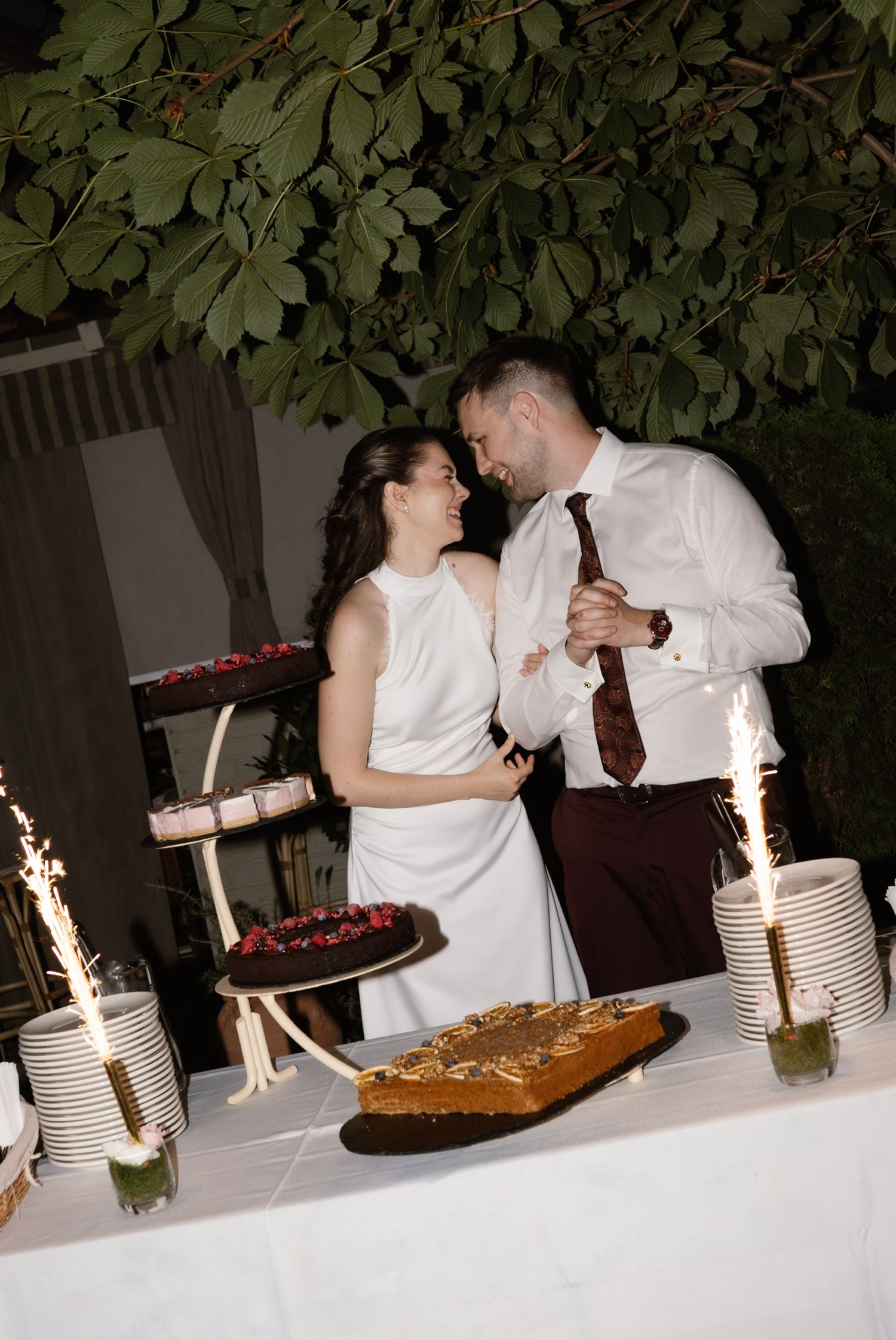 Newlyweds laughing together at wedding cake table surrounded by sparklers at night.